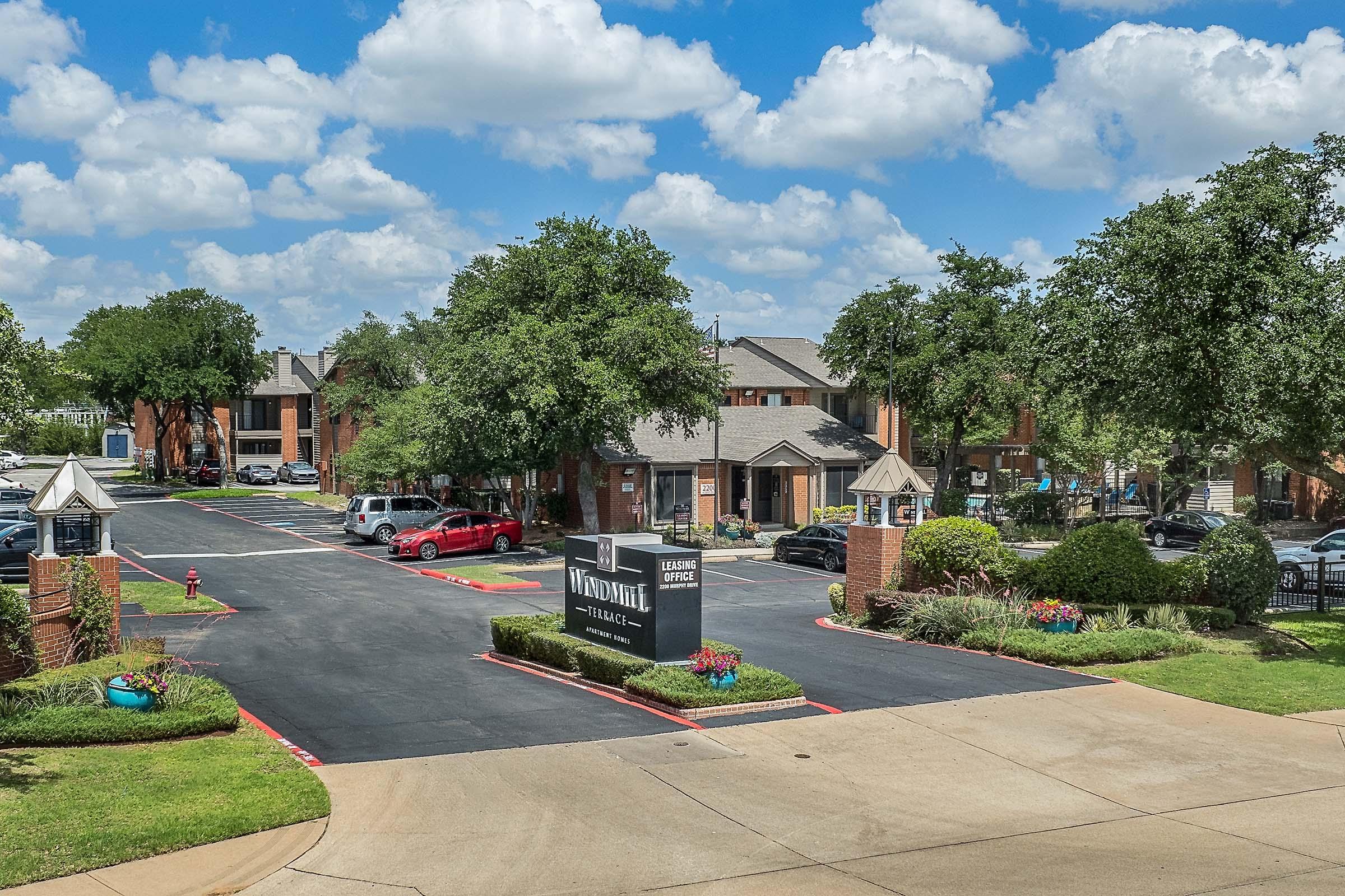 View of a residential apartment complex featuring multiple buildings with bright red brick facades, landscaped areas, and a sign for the leasing office. The parking lot is visible with several parked vehicles, and the scene is set against a blue sky with fluffy clouds.