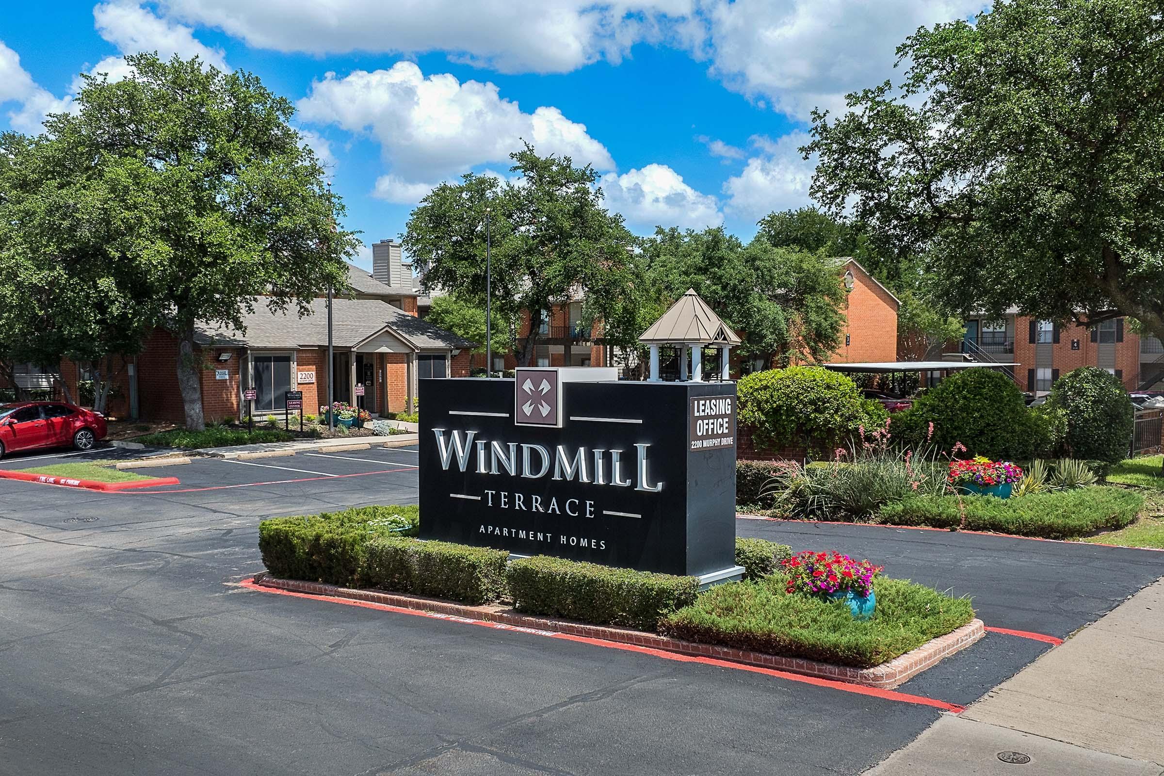 Sign for Windmill Terrace Apartment Homes, featuring the name prominently displayed. The area includes well-maintained landscaping, green trees, and a gazebo. Nearby parking is visible, along with a red car and several apartment buildings in the background under a partly cloudy sky.