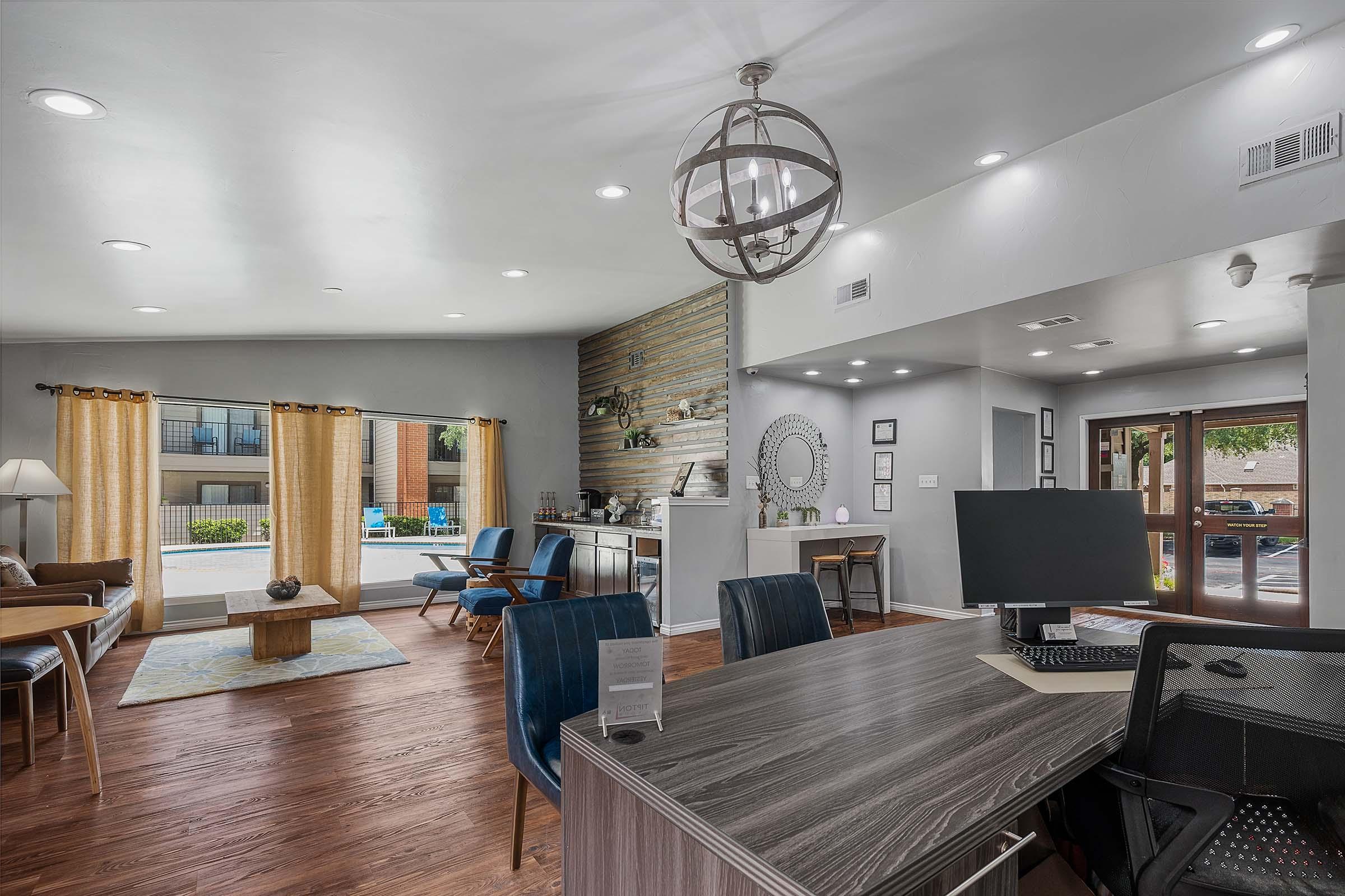 A modern lobby area with wooden flooring and large windows. There are comfortable seating options, a round chandelier, and a desk with a computer. Decorative elements like a mirror and artwork are visible. Natural light fills the space, creating a welcoming atmosphere.
