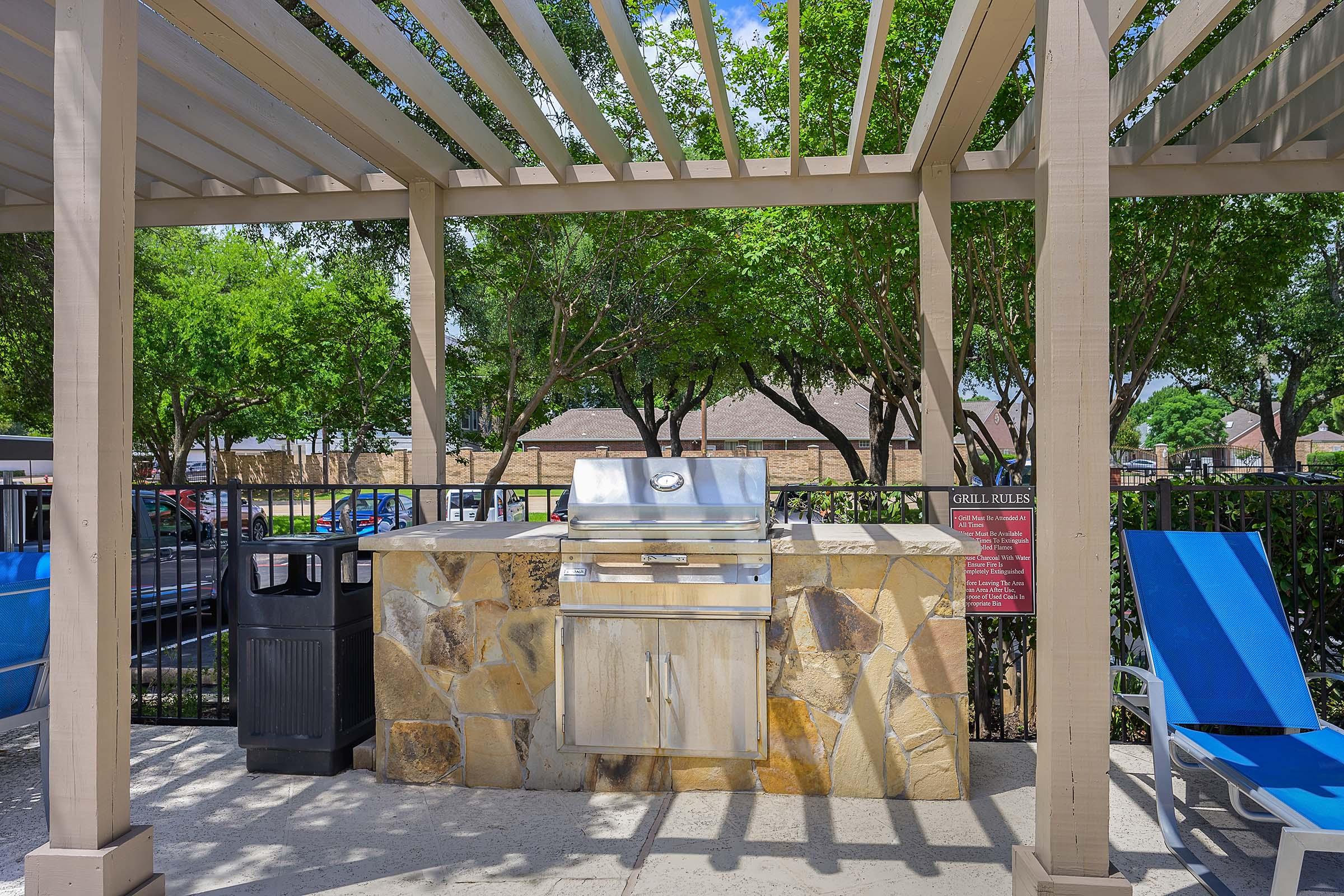 Outdoor grilling area with a stone barbecue grill, a trash can nearby, and blue lounge chairs. Surrounded by a pergola and trees, the space is well-lit with plenty of natural light. A sign with grill rules is visible on the side.