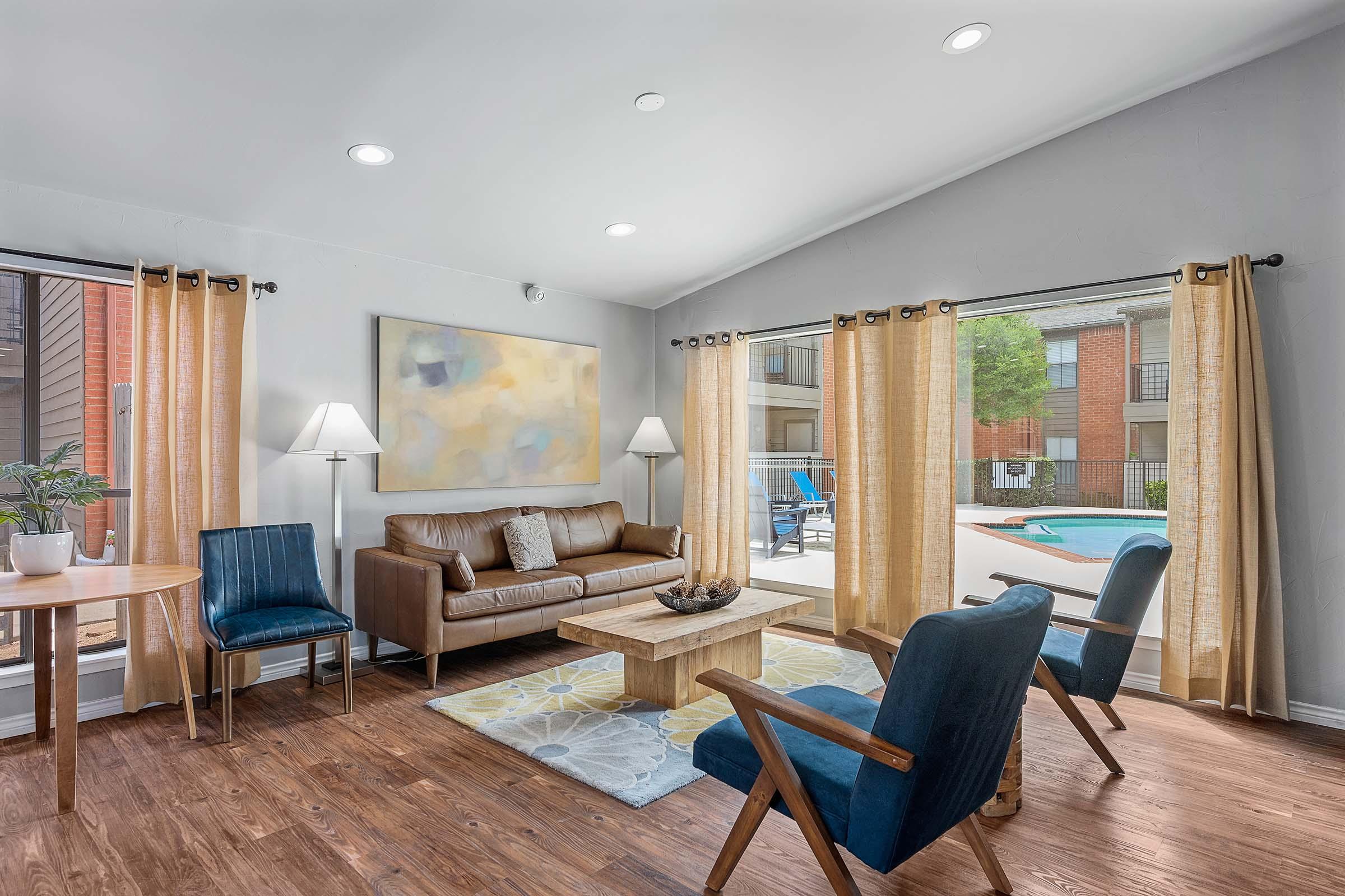 A spacious, modern living room featuring a brown leather sofa, a wooden coffee table, and two blue chairs. Large windows with sheer curtains allow natural light, revealing a view of a swimming pool outside. A small dining table and a decorative plant add to the inviting atmosphere.