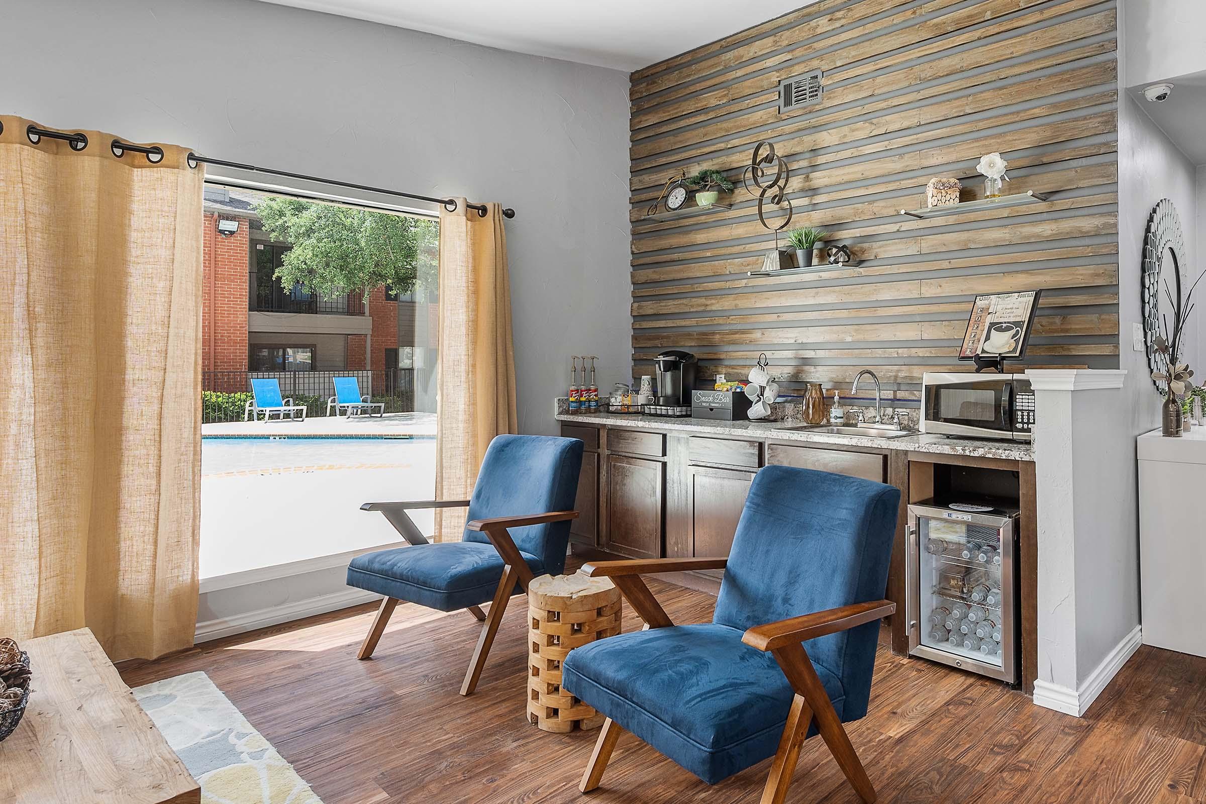 A cozy lounge area featuring two blue armchairs, a small wooden table, and a kitchenette against a wooden accent wall. Large windows offer a view of a pool area with lounge chairs outside, illuminated by natural light, creating a welcoming atmosphere.