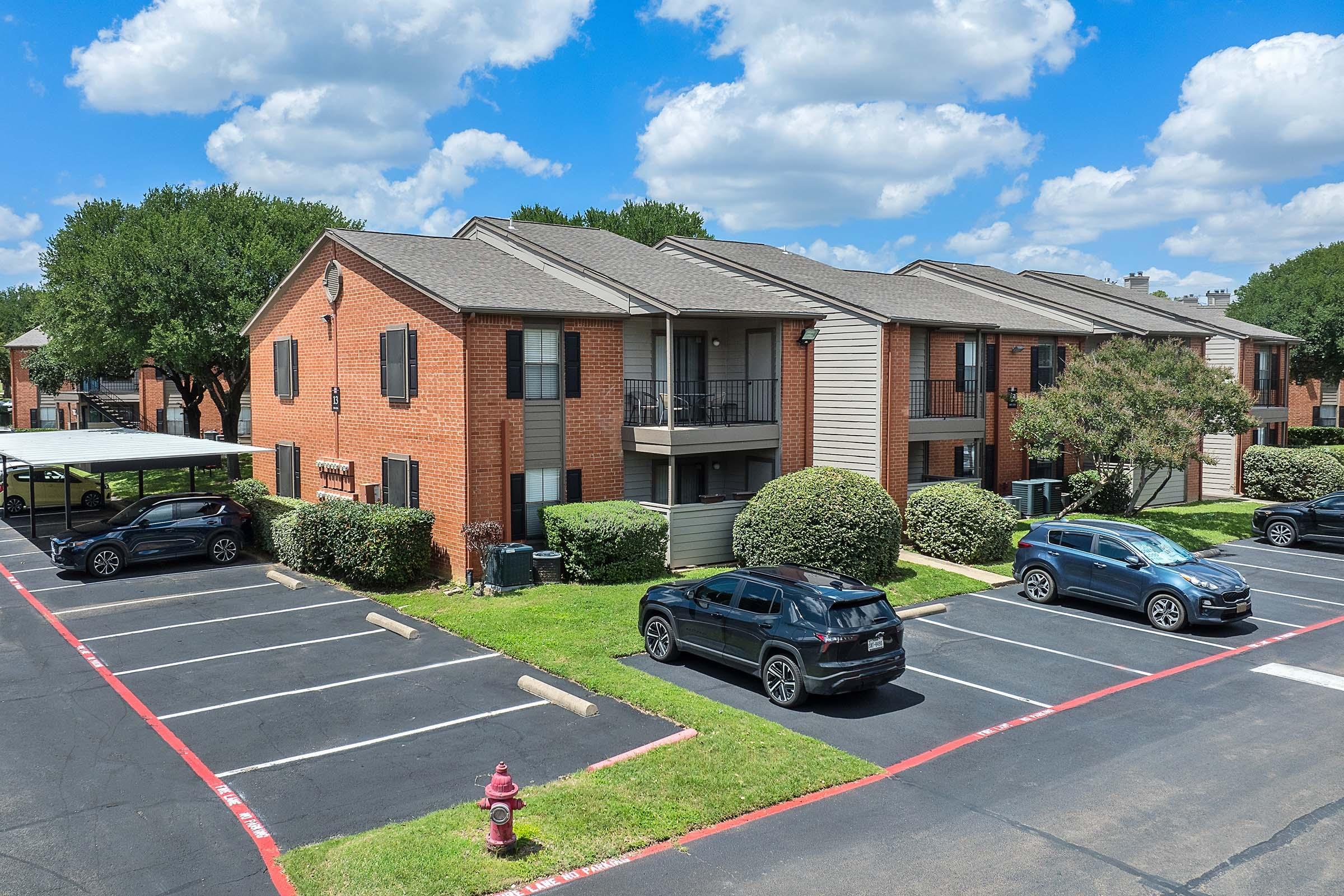 Two-story brick apartment buildings surrounded by green landscaping. Several vehicles parked on a paved lot, with a fire hydrant visible. The sky is partly cloudy, creating a bright daytime atmosphere.