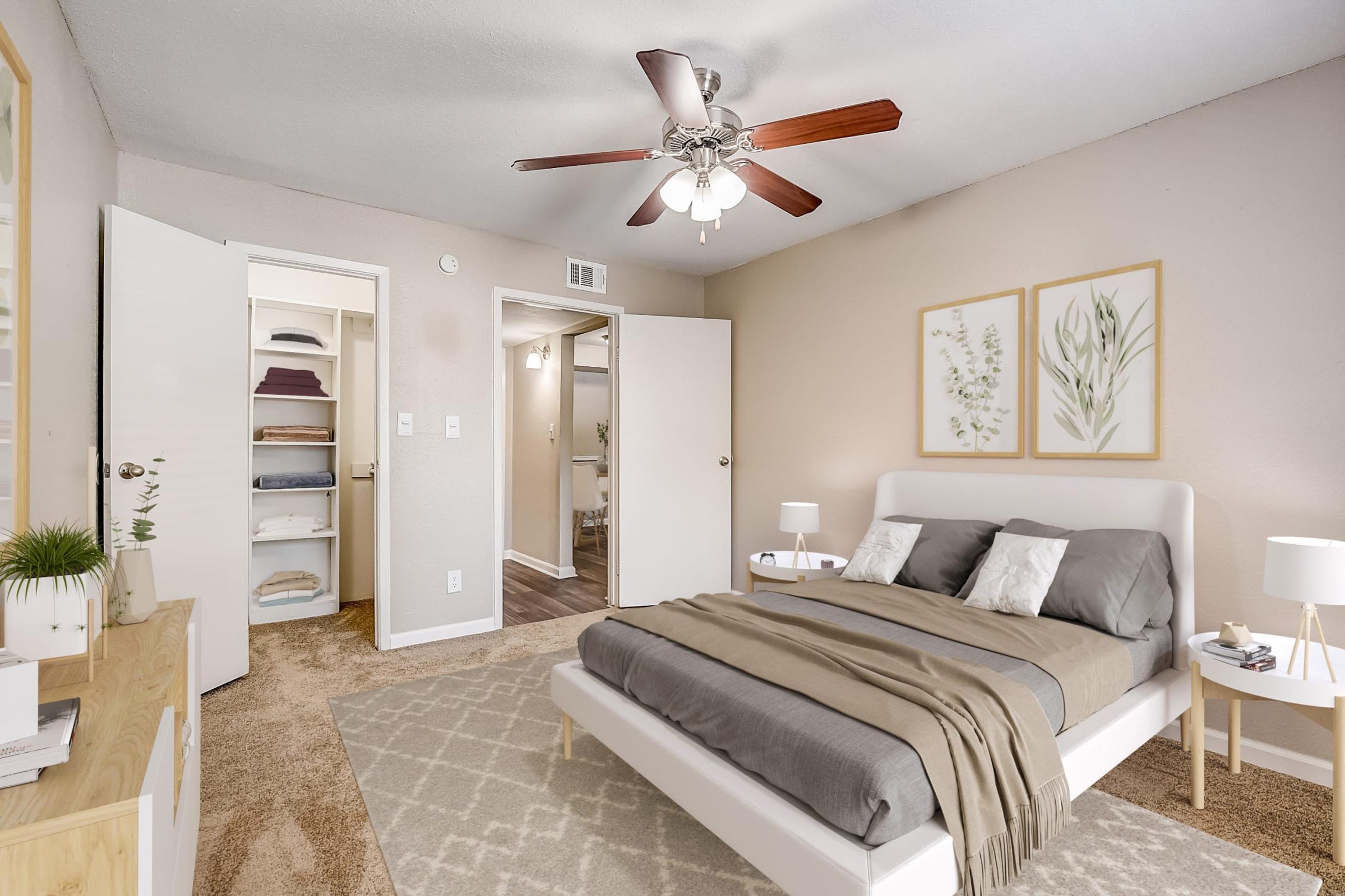 A cozy bedroom featuring a light-colored bed with gray bedding and decorative pillows. There’s a ceiling fan with wooden blades above, and light-colored walls adorned with framed botanical prints. A nightstand on either side of the bed holds small lamps, and a closet is visible in the background. The room has warm, inviting tones.