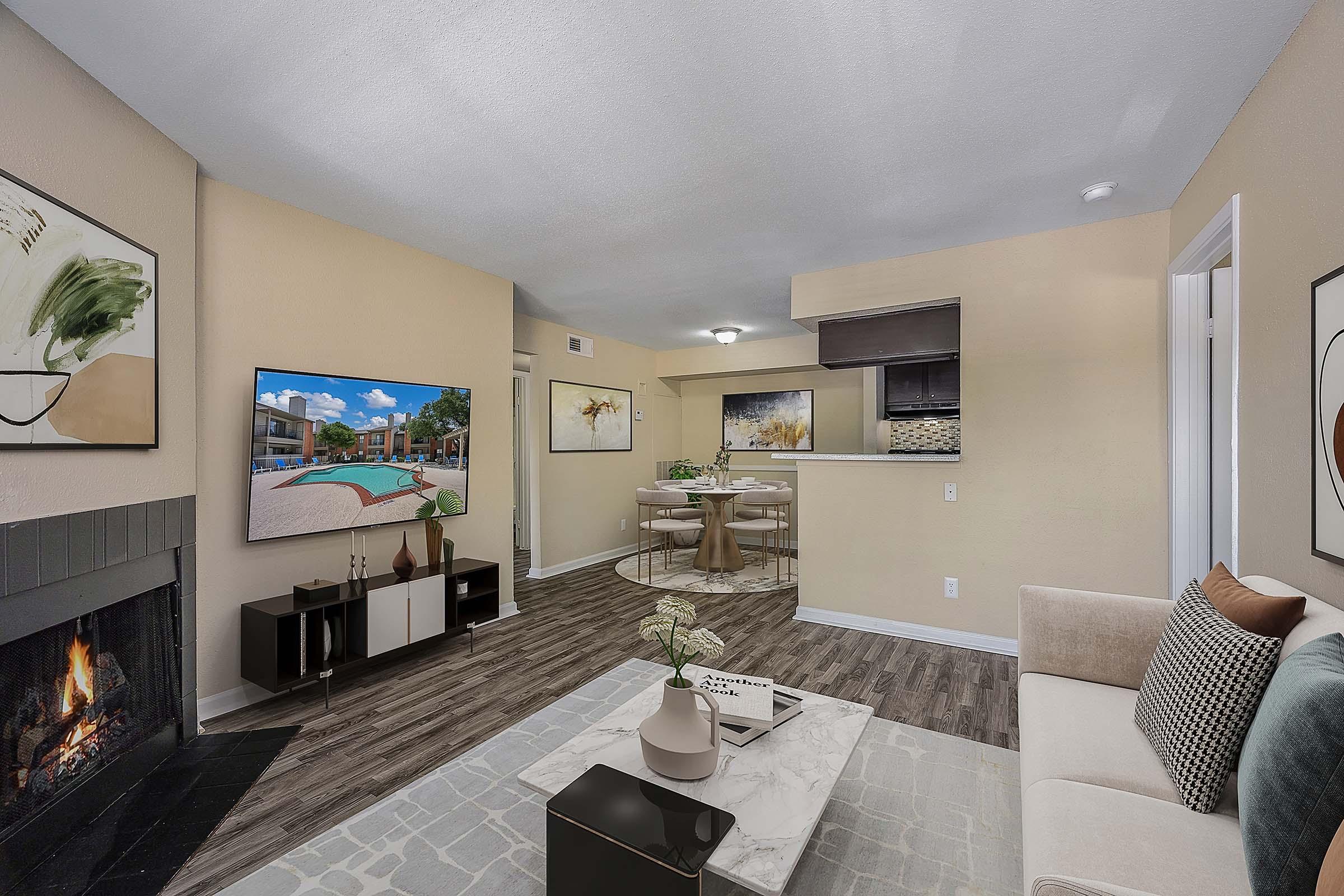 A cozy and modern living room featuring a beige wall with a large flat-screen TV displaying a pool scene. There’s a stylish coffee table on a textured rug, a comfortable sofa adorned with pillows, and a dining area in the background with a round table and tasteful decor. A fireplace adds warmth to the space.