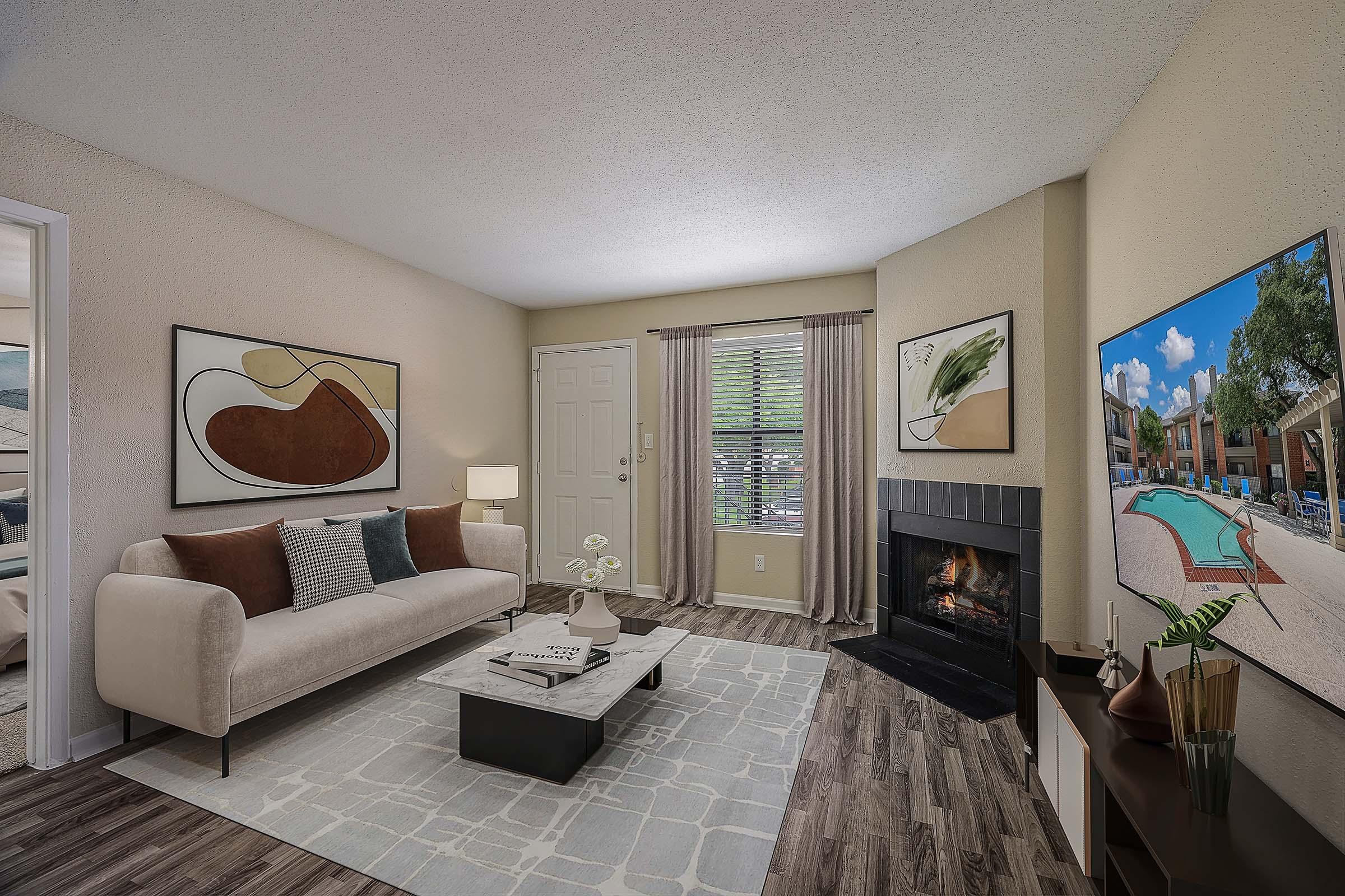 A cozy living room featuring a light-colored sofa with decorative pillows, a coffee table with magazines, and modern artwork on the walls. A television displays an outdoor pool scene. Natural light filters through window blinds, and there is a fireplace creating a warm atmosphere.