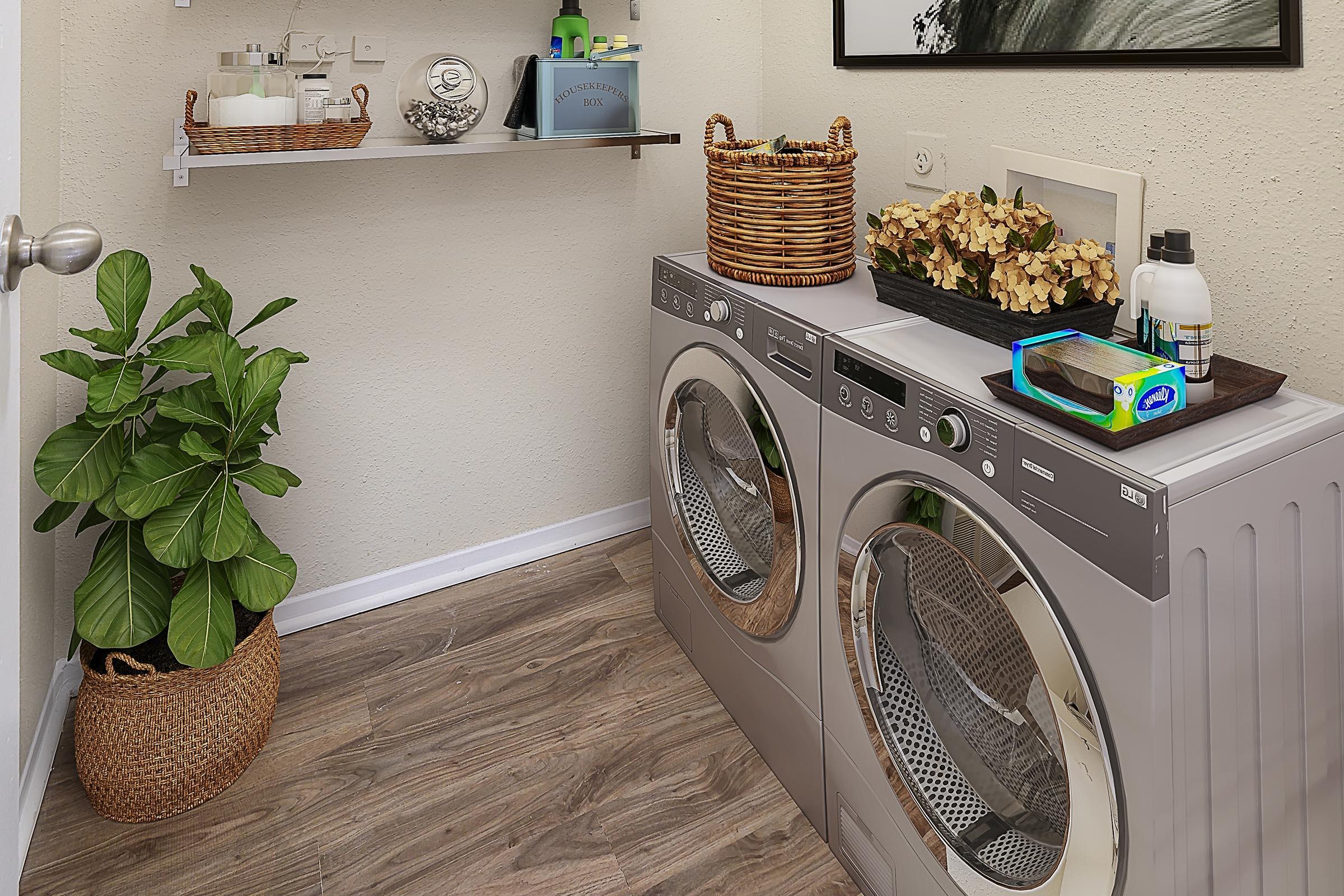 A modern laundry room featuring silver front-loading washing machine and dryer side by side. On the left, there is a potted plant in a woven basket. Above the machines, a shelf holds decorative items and storage containers. The floor has wood-like laminate, and the walls are painted a light color.