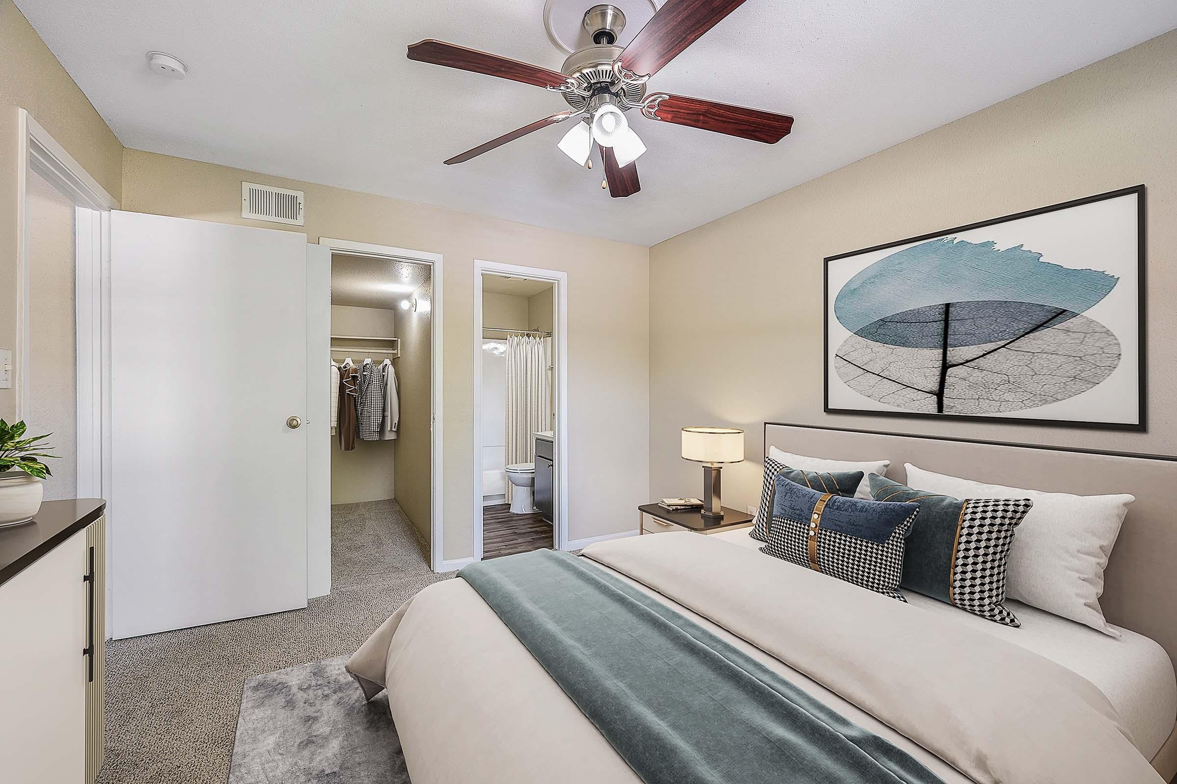 A cozy bedroom featuring a bed with a soft grey and blue comforter, decorative pillows, and a wall-mounted artwork. A bedside lamp adds warmth, while a ceiling fan provides airflow. The room leads to a closet and a bathroom area visible through an open door.
