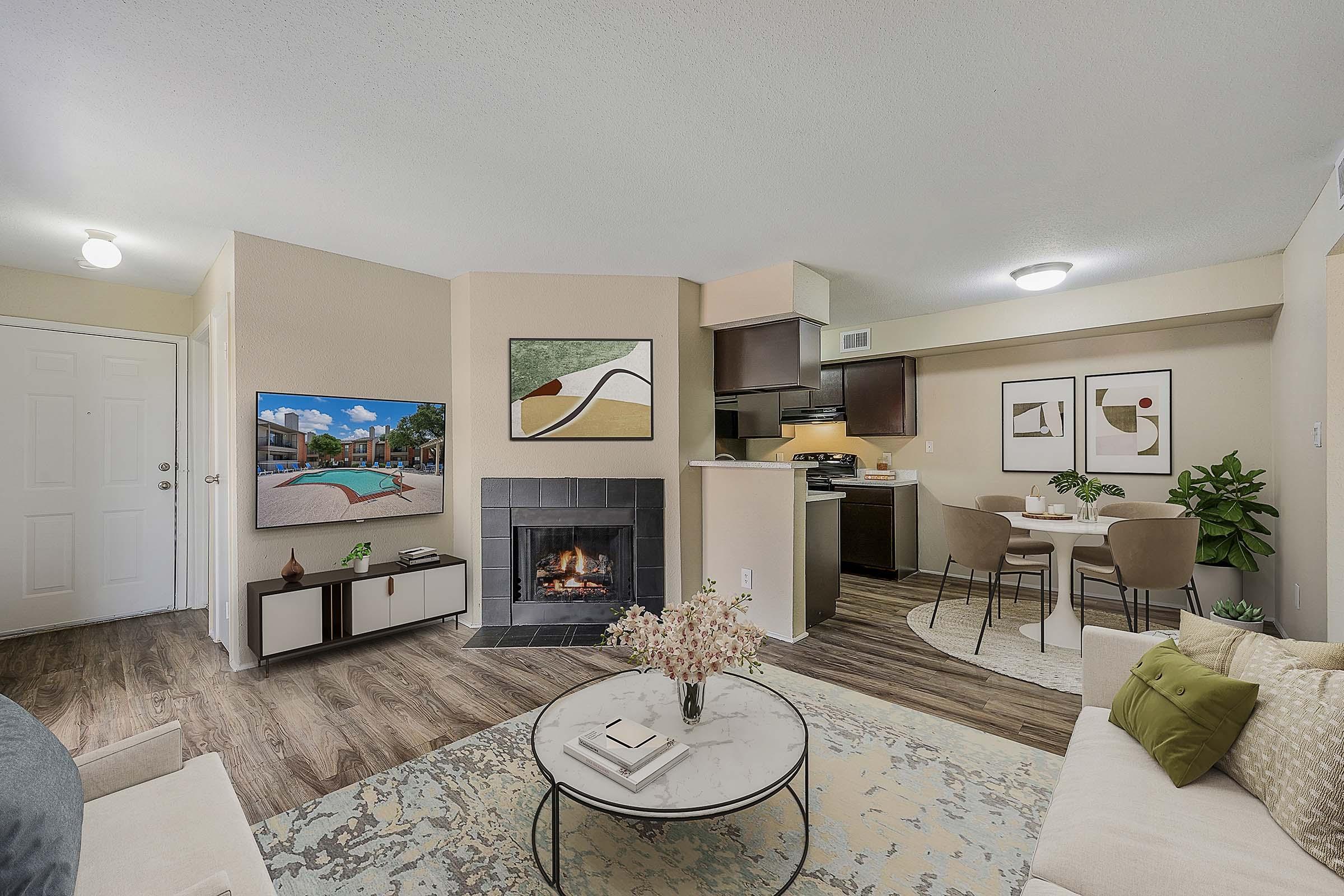 A cozy living room featuring a modern design with a white sofa, a round coffee table, and a fireplace. There are framed artworks on the walls, a TV mounted above the fireplace, and a dining area with chairs. The space has wooden flooring and plants for a fresh touch.