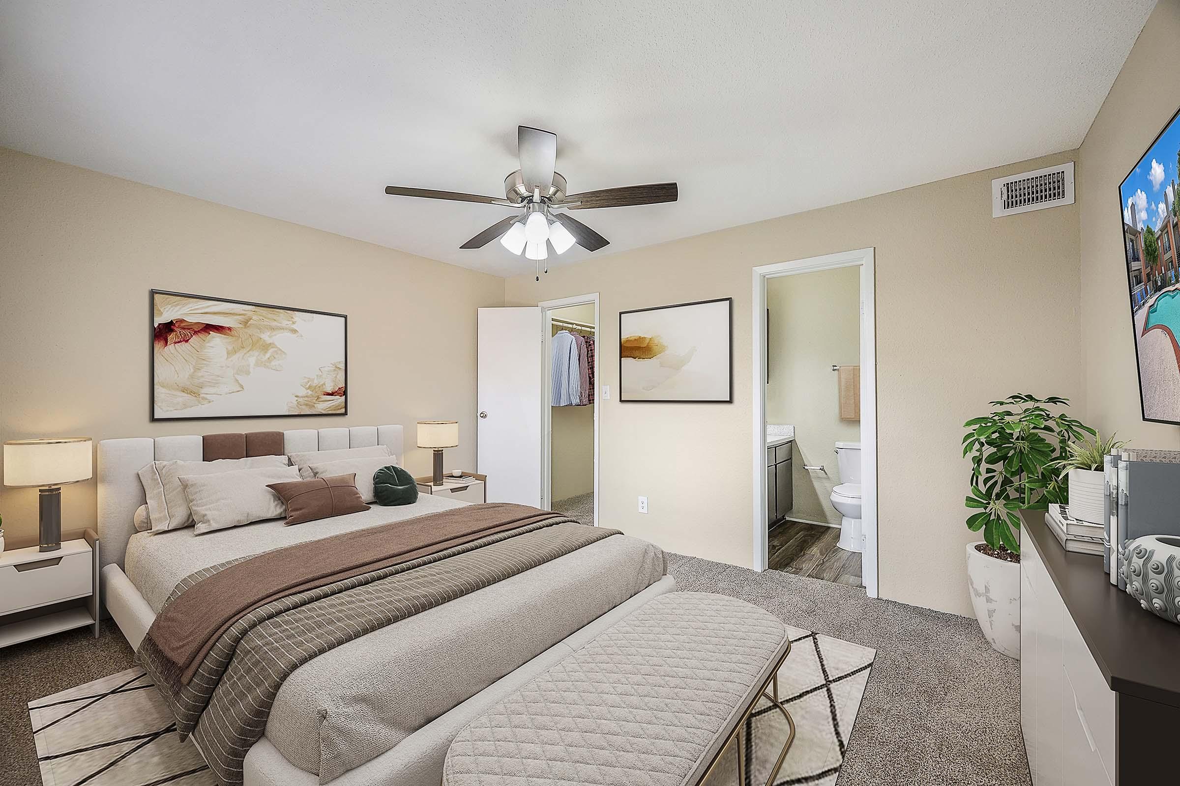 A cozy bedroom featuring a queen-sized bed with neutral bedding, two bedside lamps, and wall art. There's a ceiling fan above and a potted plant in the corner. A door leads to a closet, while another door opens to a bathroom. The room has light beige walls and a comfortable, inviting atmosphere.