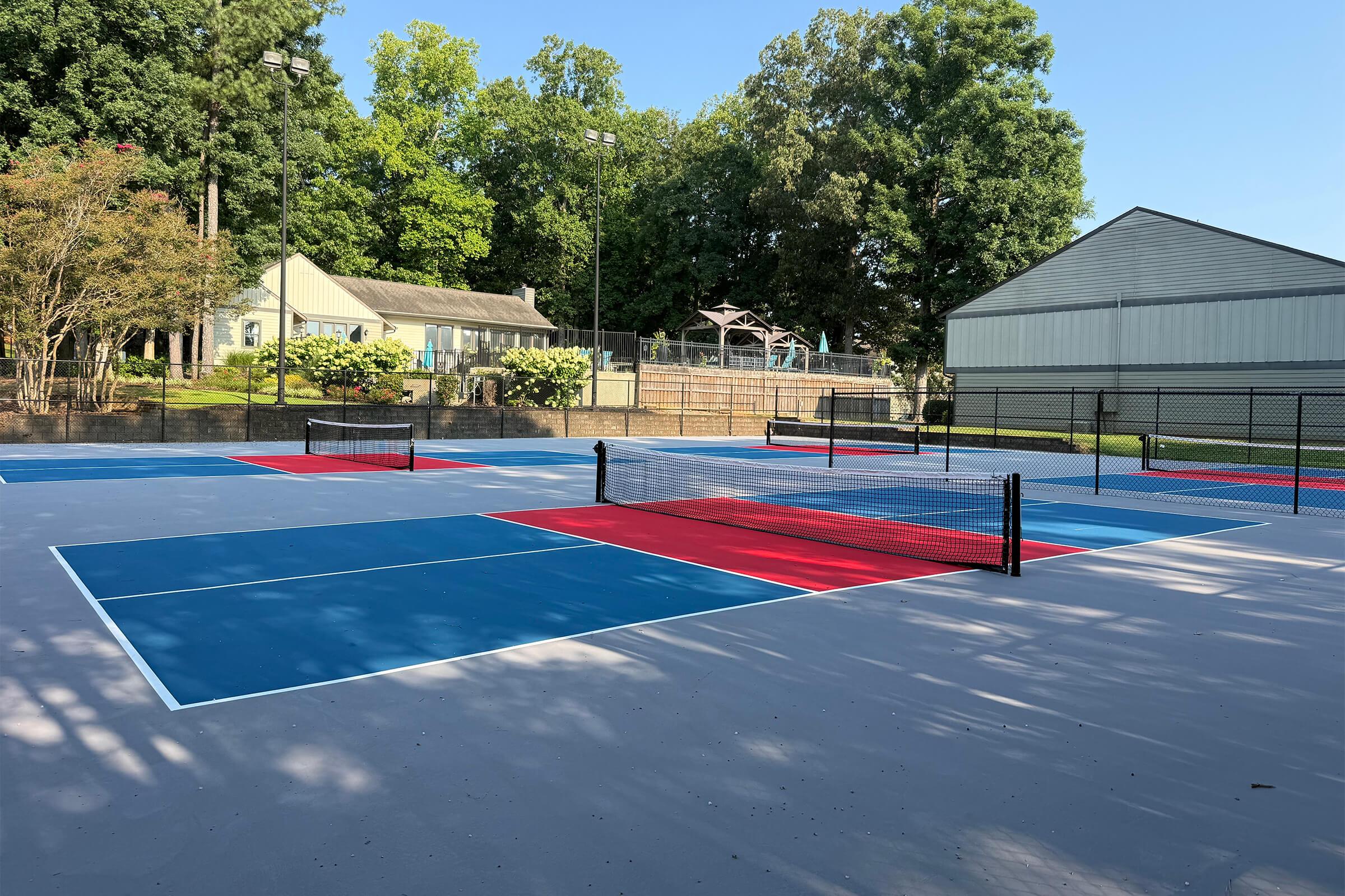 A brightly colored outdoor tennis court featuring blue and red sections, surrounded by trees. In the background, there is a playground and a large building. The scene is well-lit, suggesting a sunny day, with clear lines marking the playing areas and nets in place. Ideal for recreational tennis play.