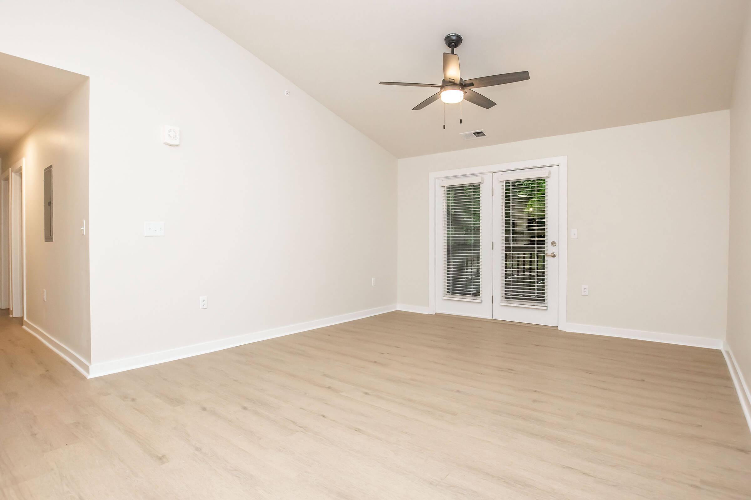 A spacious, empty room with light-colored walls and wooden flooring. It features a ceiling fan and large double doors leading to an outdoor area, with natural light coming in through the windows. The room has a simple design, suitable for various decorating styles.