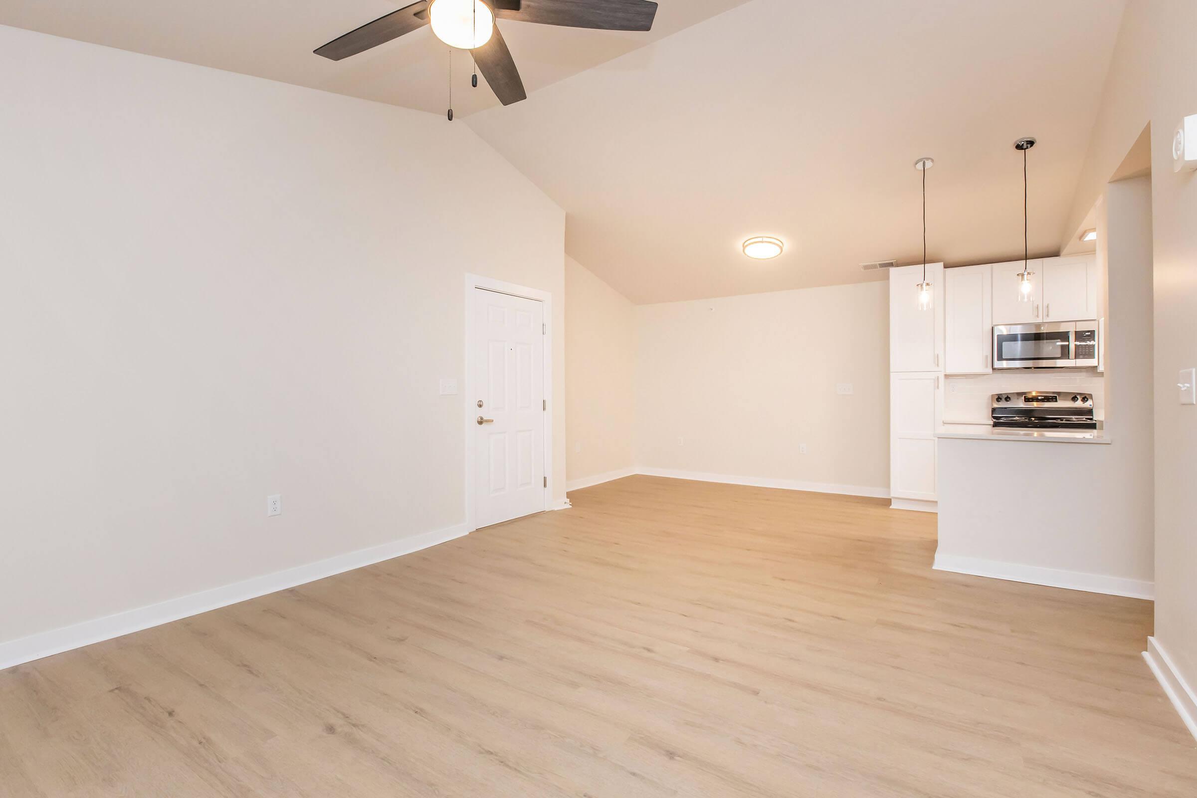 A spacious, modern living area featuring light-colored walls, a ceiling fan, and large windows. There’s a kitchen with stainless steel appliances and white cabinetry visible in the background. The floor is light wood, and the overall design is open and inviting.
