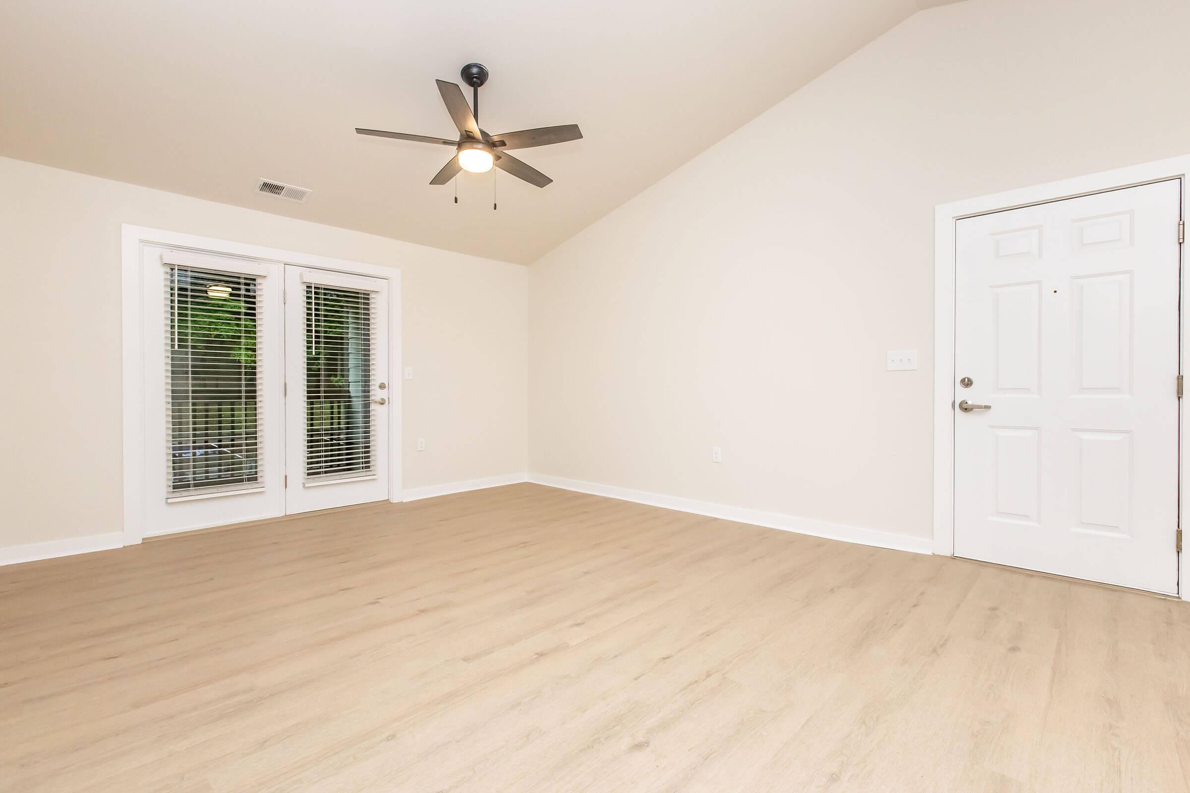 Bright and spacious room featuring light-colored walls, hardwood floors, and a ceiling fan. Large windows with blinds provide natural light, and a white door offers access to the outside. The room has a simple, modern aesthetic, making it versatile for various uses.