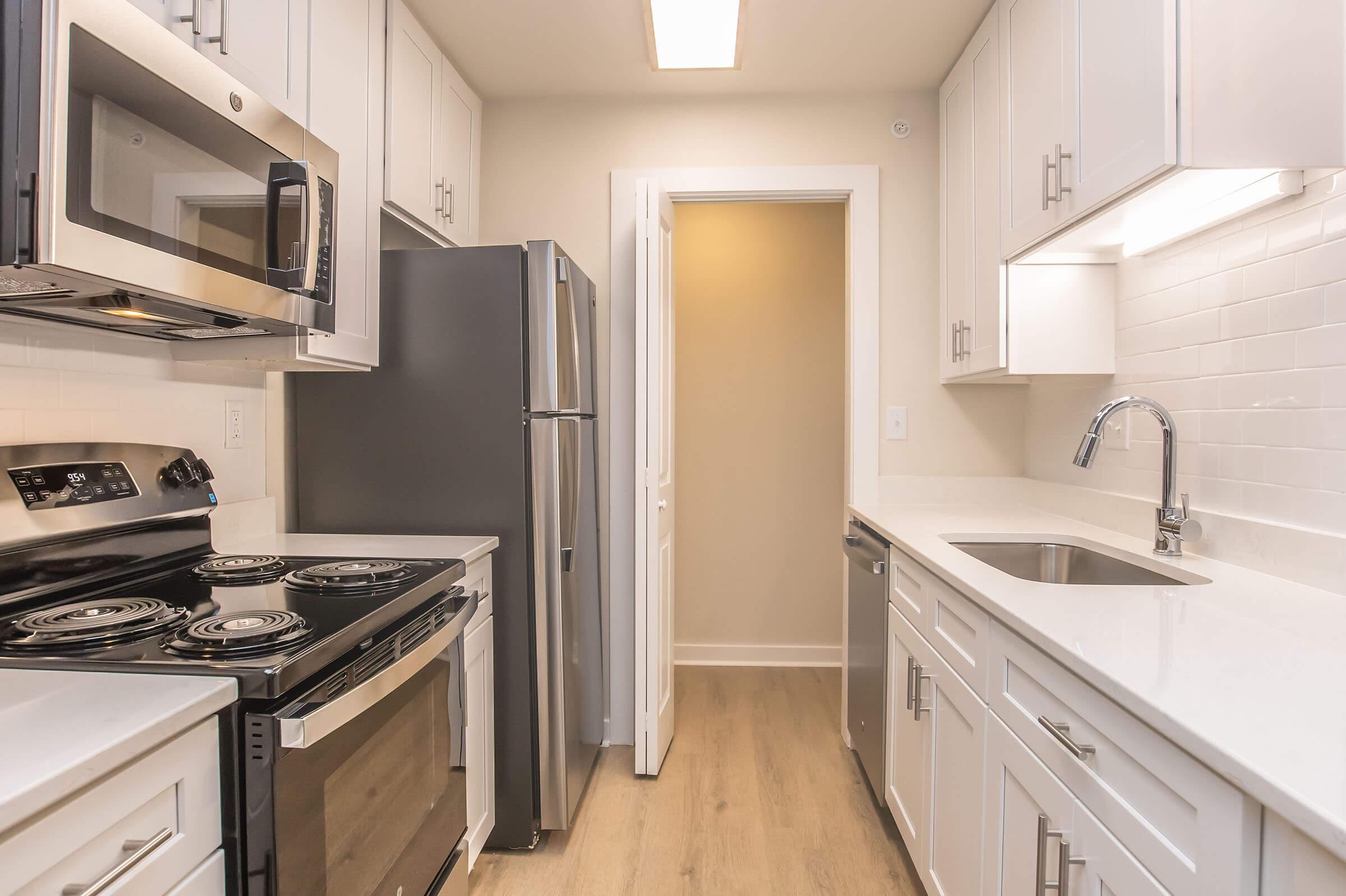 Modern kitchen featuring stainless steel appliances, including a microwave and refrigerator. There is a stove with four burners, a sleek sink with a pull-down faucet, and white cabinets. Natural light filters in from an overhead fixture, enhancing the clean, contemporary design with light wood flooring.