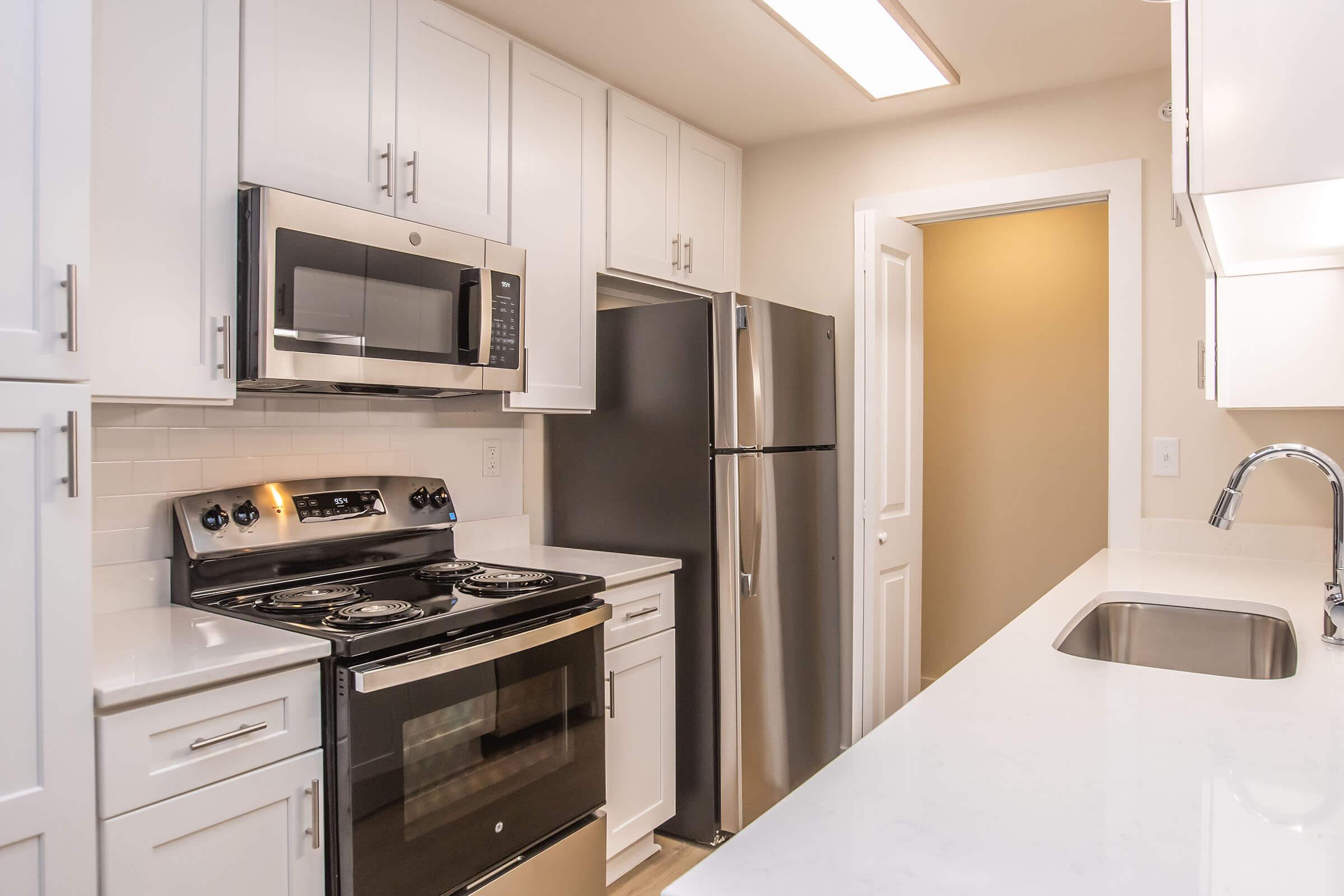 Modern kitchen featuring white cabinetry, stainless steel appliances including a microwave, oven, and refrigerator. The countertop is light-colored, with a sink visible. The kitchen has an open layout, and a door leads to another room. Bright lighting enhances the overall spacious feel.