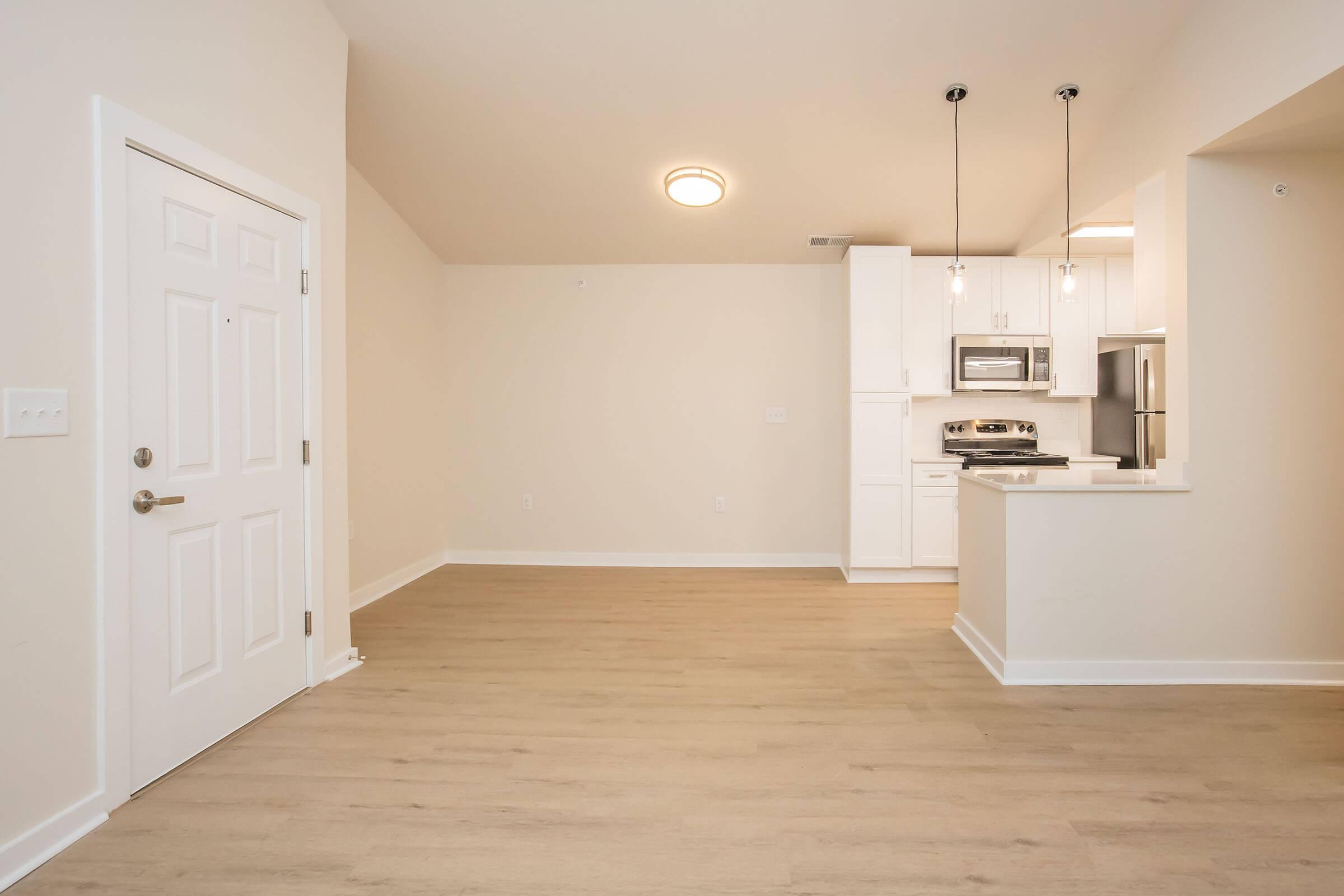 A bright and spacious living area featuring light-colored walls and a neutral floor. In the background, a modern kitchen with white cabinets, stainless steel appliances, and pendant lighting is visible. The entrance door is on the left, enhancing the open feel of the space.