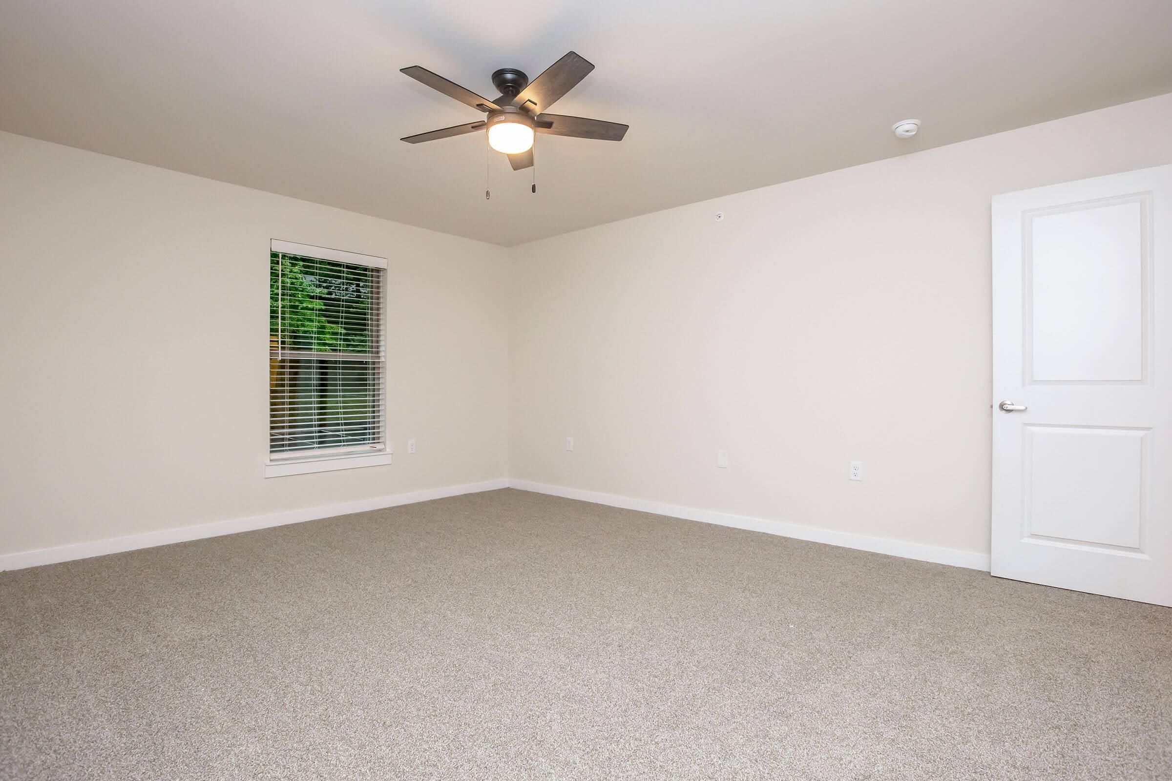 A spacious, empty room with beige walls and carpeted flooring. A ceiling fan with black blades is mounted above, and there is a single window with white blinds letting in natural light. A white door leads to another space, creating a clean and minimalistic atmosphere.