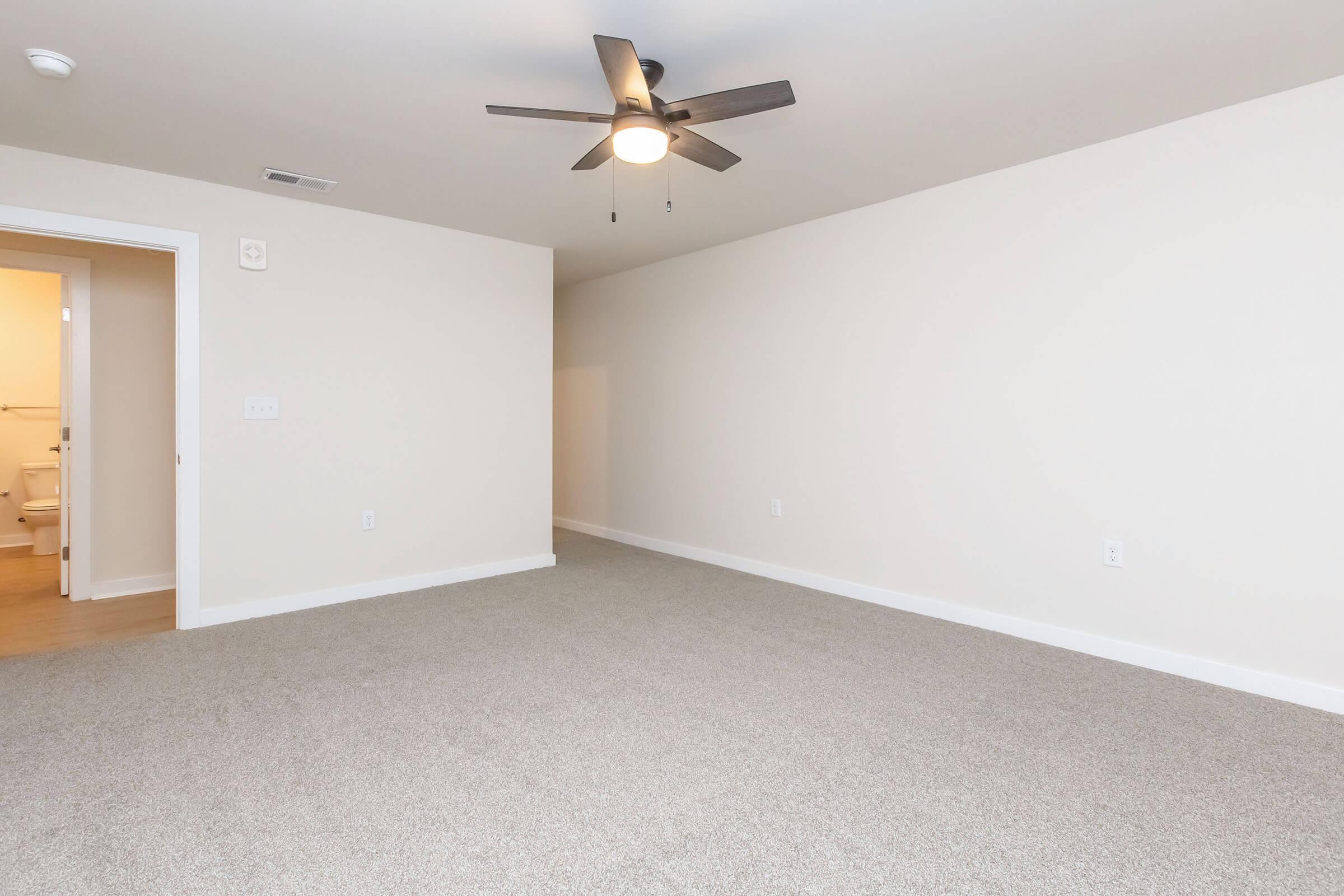 A spacious, unfurnished room featuring light beige walls and a ceiling fan. The floor is covered in light gray carpeting. To the left, there is a doorway leading to a bathroom. The overall atmosphere is bright and airy, with natural light coming in from the doorway.