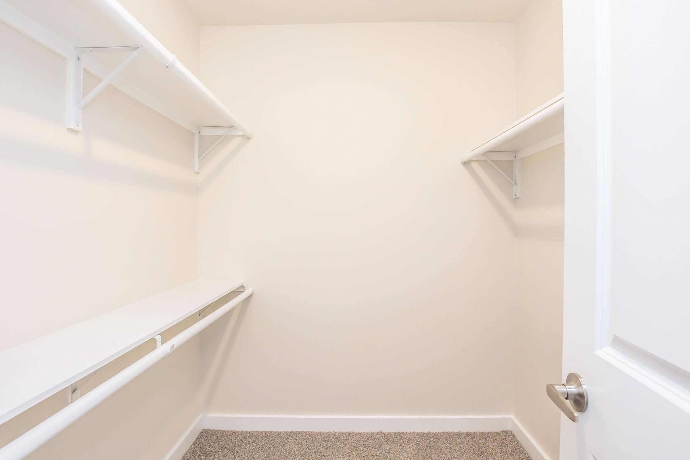 An empty walk-in closet featuring beige walls and light gray carpeting. Two white shelves run along three walls, with a door to the right. The space is well-lit and clean, offering a minimalist look with no items or furnishings inside.