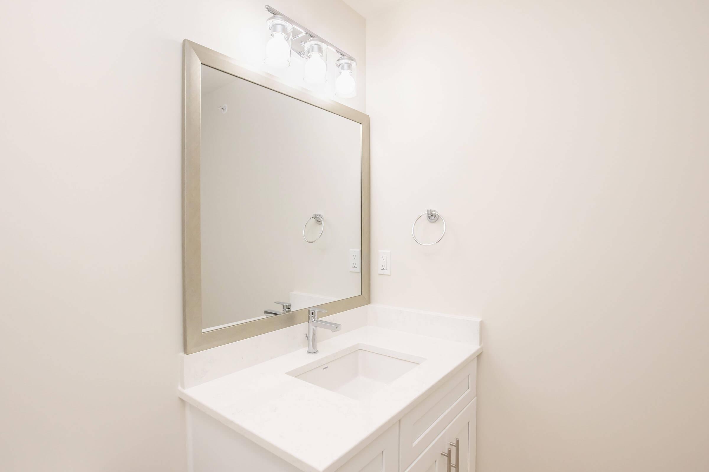 A modern bathroom vanity with a white countertop and sink. Above the sink is a large, rectangular mirror framed in a light-colored metal. There is a sleek faucet installed on the countertop. The walls are painted a soft, neutral shade, and a wall-mounted towel holder is visible beside the mirror.