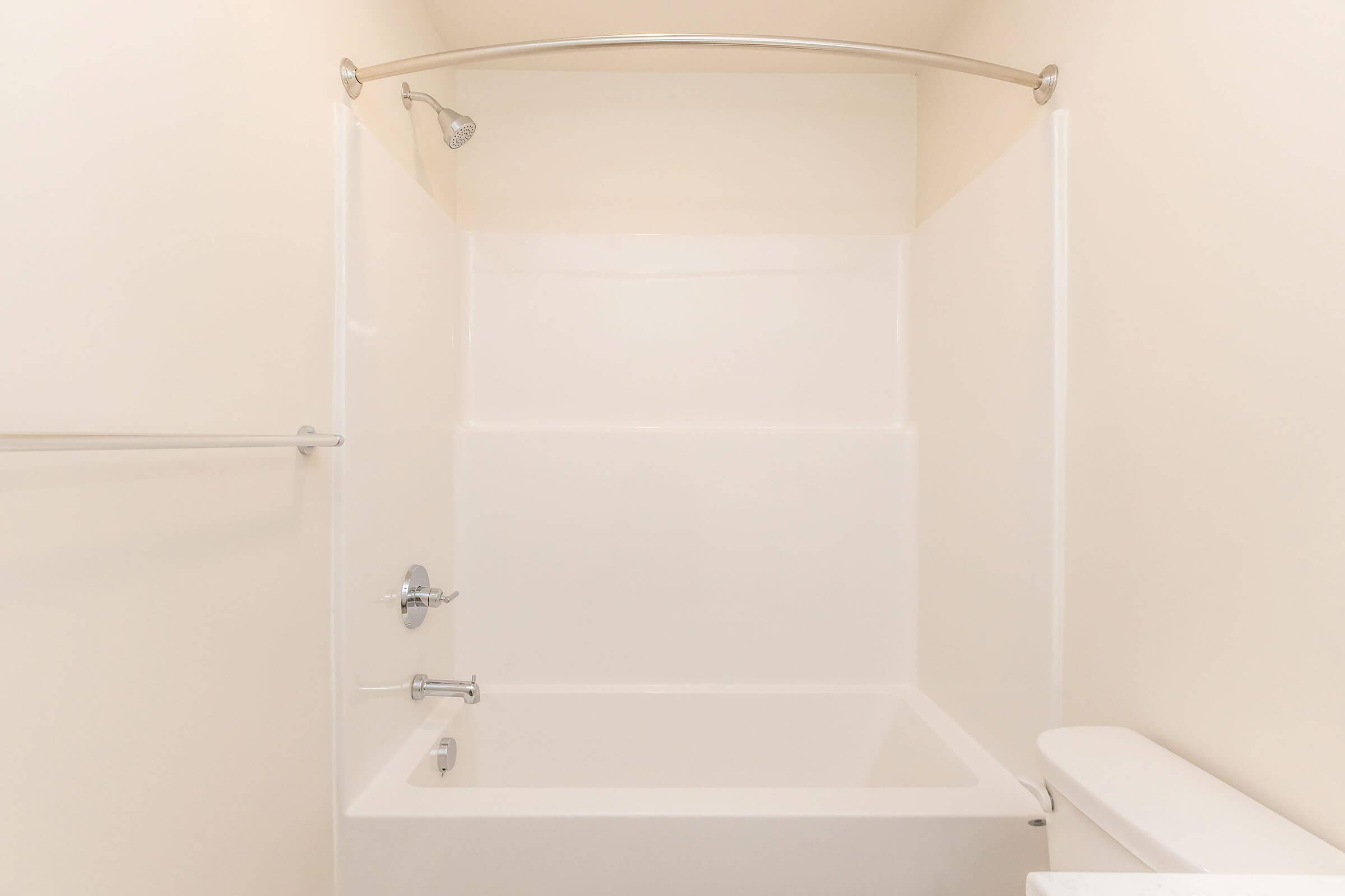 A clean, empty bathroom with a white tub and a shower head above. The walls are plain and beige, providing a simple, modern aesthetic. A towel bar is mounted on the left side, and a white toilet is visible in the lower right corner, completing the uncluttered look of the space.