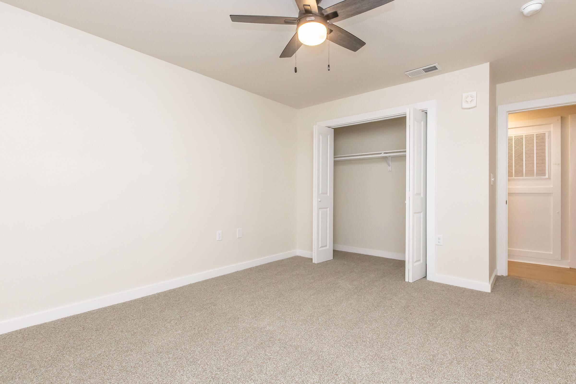A spacious, empty room with light beige walls and a ceiling fan. The floor is carpeted in a light tan shade. There is a closet with double white doors on one side, and a doorway leading to another room is visible in the background, creating a sense of openness.