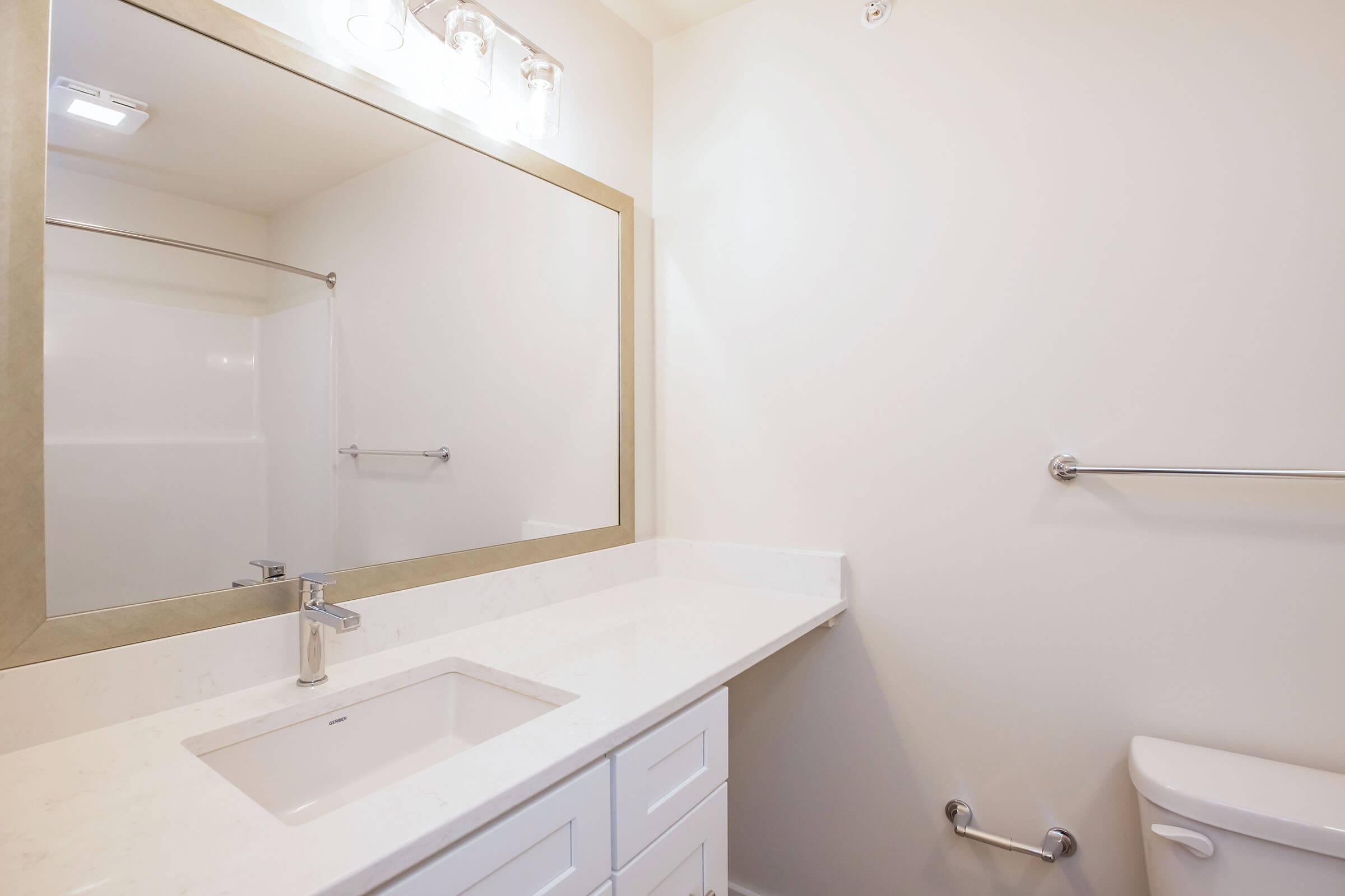 A clean and modern bathroom featuring a white countertop with a rectangular sink, a large mirror with a metallic frame, a shower area with a glass sliding door, and a toilet. The walls are painted a light color, and there is a towel bar beside the sink. Bright lighting enhances the spacious feel.