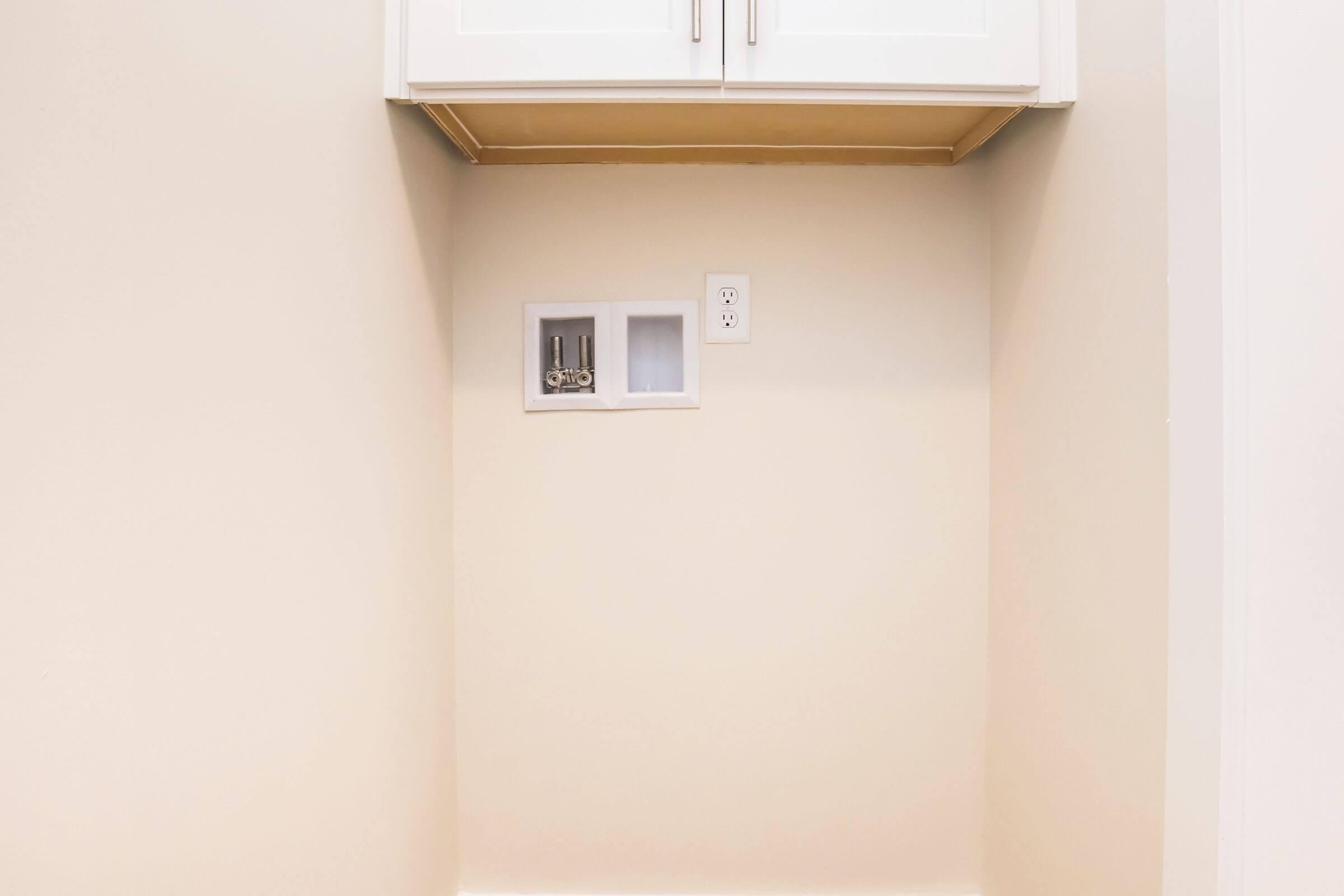 Empty wall space featuring a cabinet above and a small niche below with two gray pipes visible. To the right, there is an electrical outlet. The surrounding wall is painted a light color, creating a clean and simple appearance.