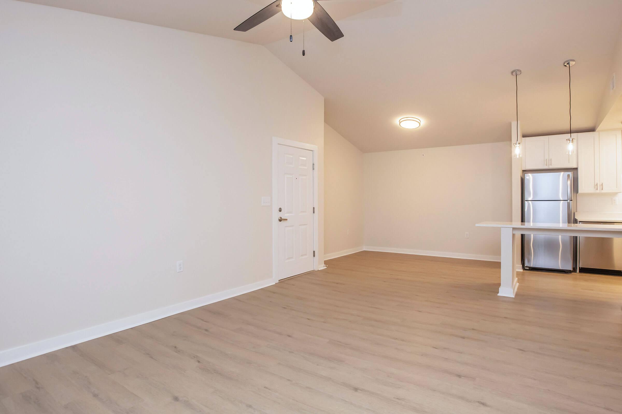 A spacious, empty room with light-colored walls and a high ceiling. A ceiling fan is installed, and there are two kitchen bar stools next to a kitchen counter. To the right, a stainless steel refrigerator is visible, along with modern cabinetry. The floor features light wood laminate, creating a bright atmosphere.