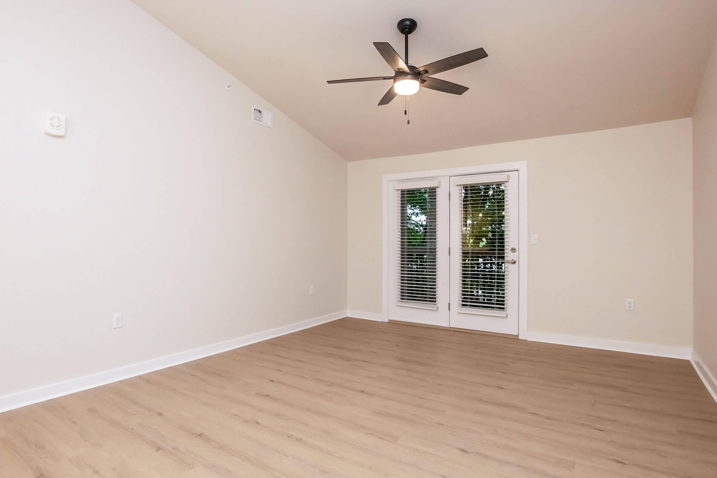 Empty room featuring light wood flooring, a ceiling fan, and large windows with blinds. The walls are painted in a light color, and there are double doors leading outside. The space has a clean and modern look, suitable for various uses or decor.