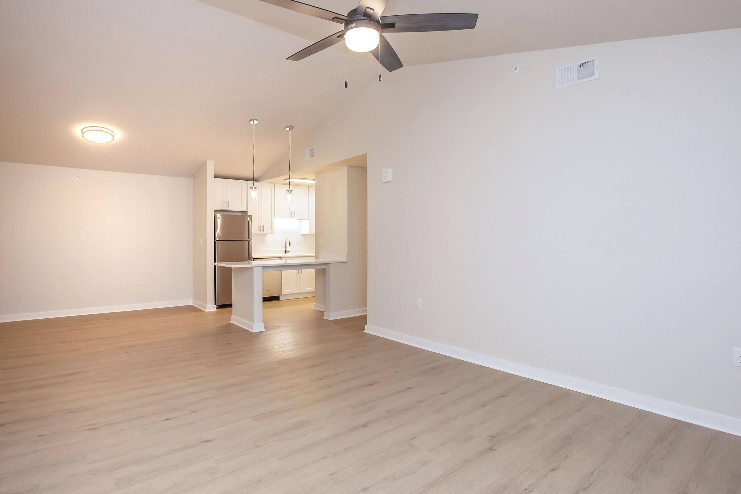 Spacious, modern interior of an apartment featuring a light-colored wall, wooden floor, and ceiling fan. The open layout connects the living area to a kitchen with white cabinets and appliances in the background. Bright, natural light flows through the space, creating an inviting atmosphere.