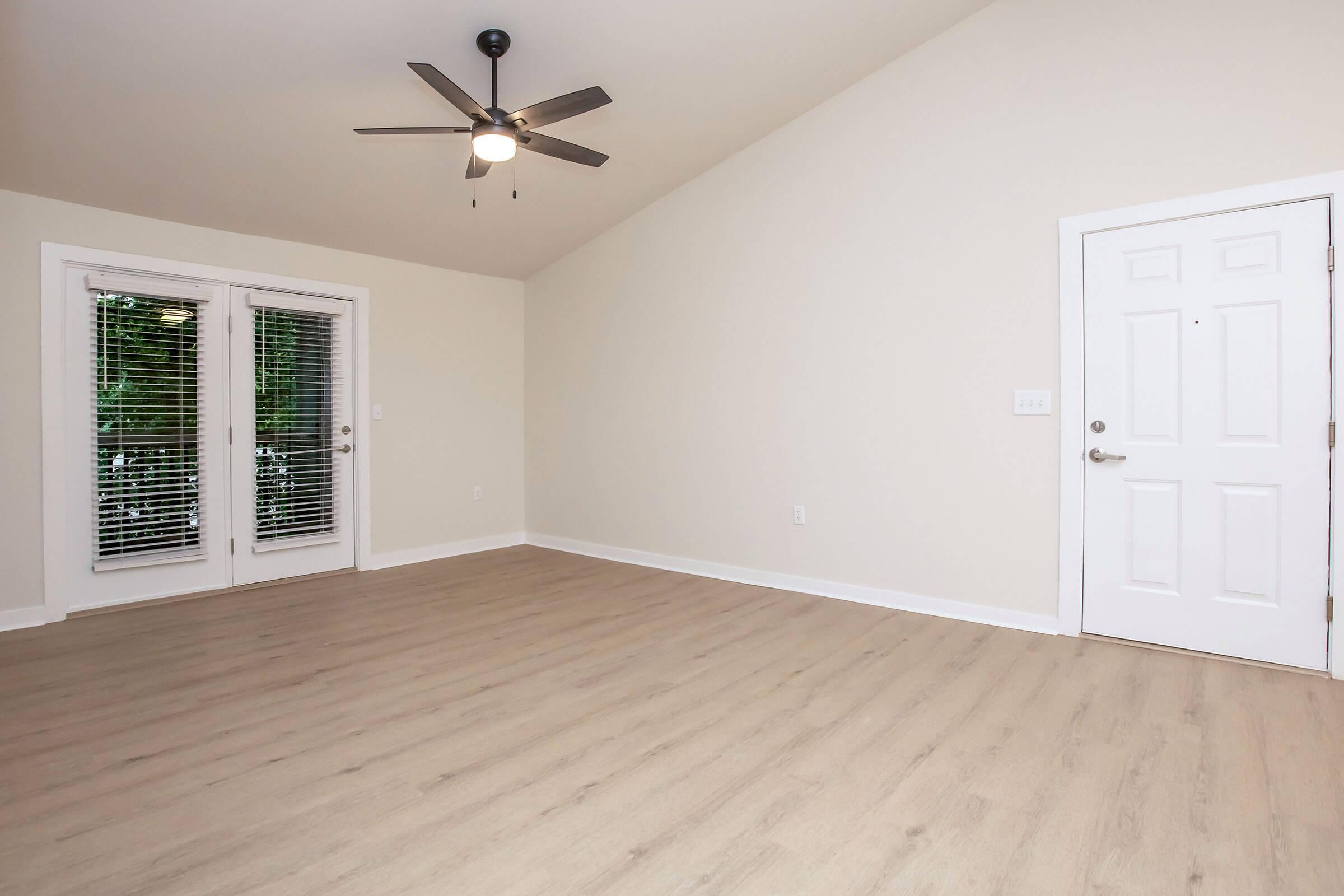 Spacious, empty room with light-colored walls and wooden flooring. A ceiling fan is mounted in the center, and there are double French doors leading to the outside. The main entry door is visible on the right, and large windows provide natural light. The room has a vaulted ceiling, creating an airy feel.