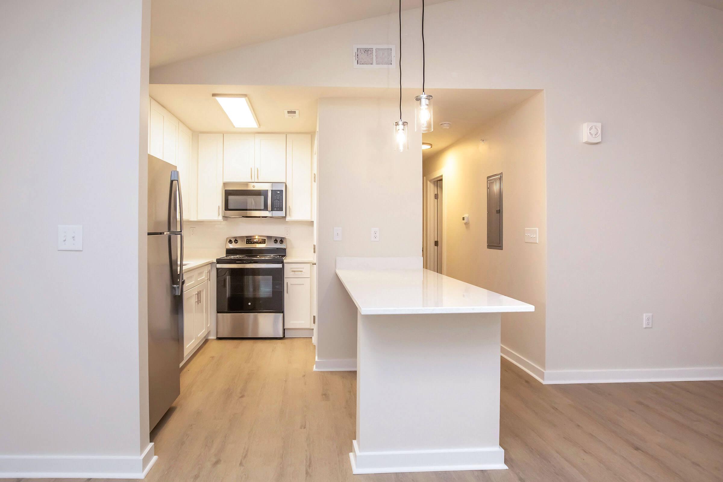 Modern kitchen with stainless steel appliances, including a refrigerator, microwave, and stove. Features white cabinetry and a large island with a countertop, pendant lighting, and light-colored flooring. Open layout connecting to a brightly lit living area.