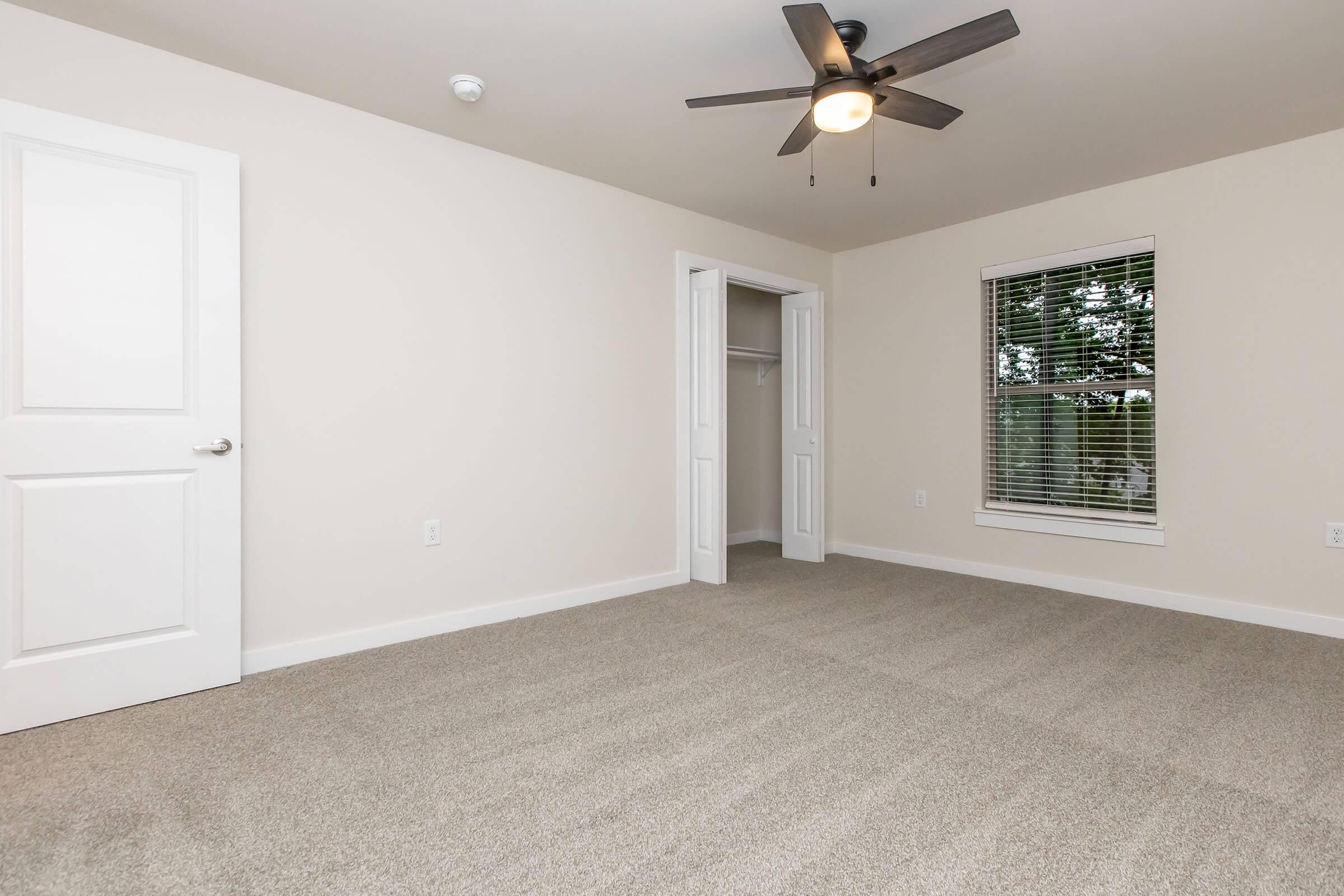 A clean, empty bedroom featuring light-colored walls, a ceiling fan, and a window with blinds. The floor is covered with light carpet, and there is a closet with double doors on one side of the room. The overall atmosphere is bright and inviting.