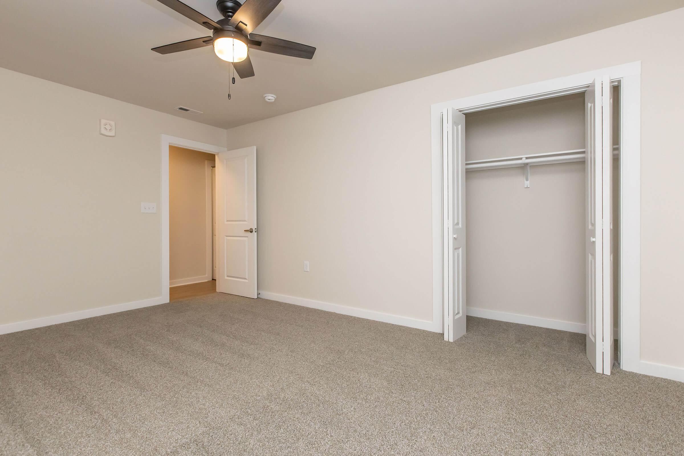 A well-lit empty room with light beige walls and carpeted flooring. It features a ceiling fan and a closet with double doors. One door leads to a hallway, providing access to the space. The overall atmosphere is spacious and ready for furniture or decoration.