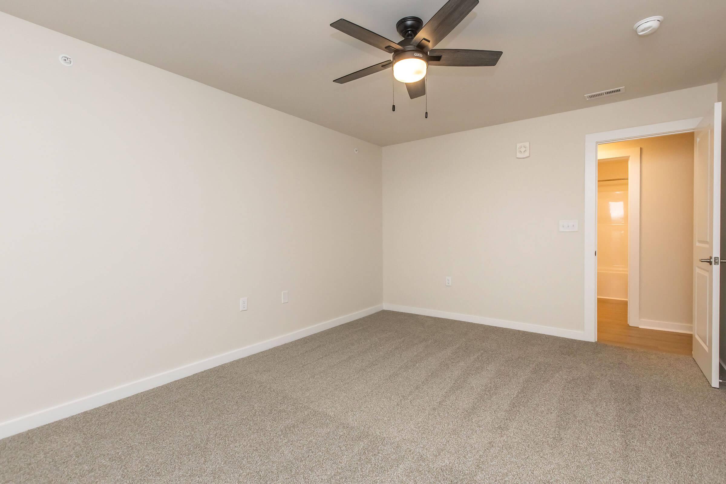 A spacious, empty room with beige walls and light brown carpet, featuring a ceiling fan with a light fixture. On one side, there is a door leading to another area, and soft lighting can be seen in the background, suggesting an adjacent area. The overall atmosphere is bright and inviting.