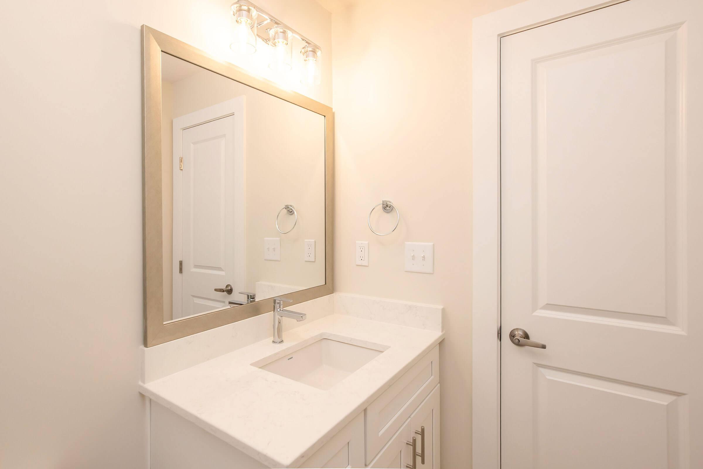 A modern bathroom featuring a white vanity with a sink and polished countertop. A large, framed mirror is above the sink, and there are two towel rings on the wall. A white door is partially visible, leading to another space. The overall color scheme is light and neutral.