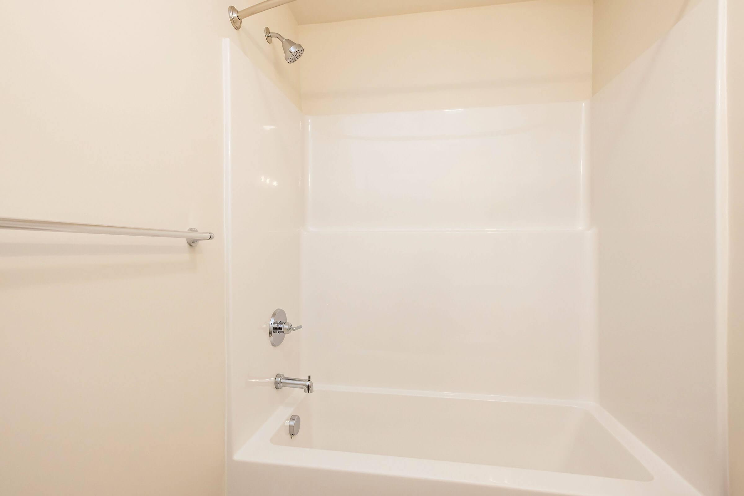 A clean and empty bathroom with a white bathtub and shower combination. The walls are light-colored, and there is a showerhead and faucet on the wall. A towel bar is mounted on the side. The space is well-lit and uncluttered, emphasizing its simplicity and modern design.