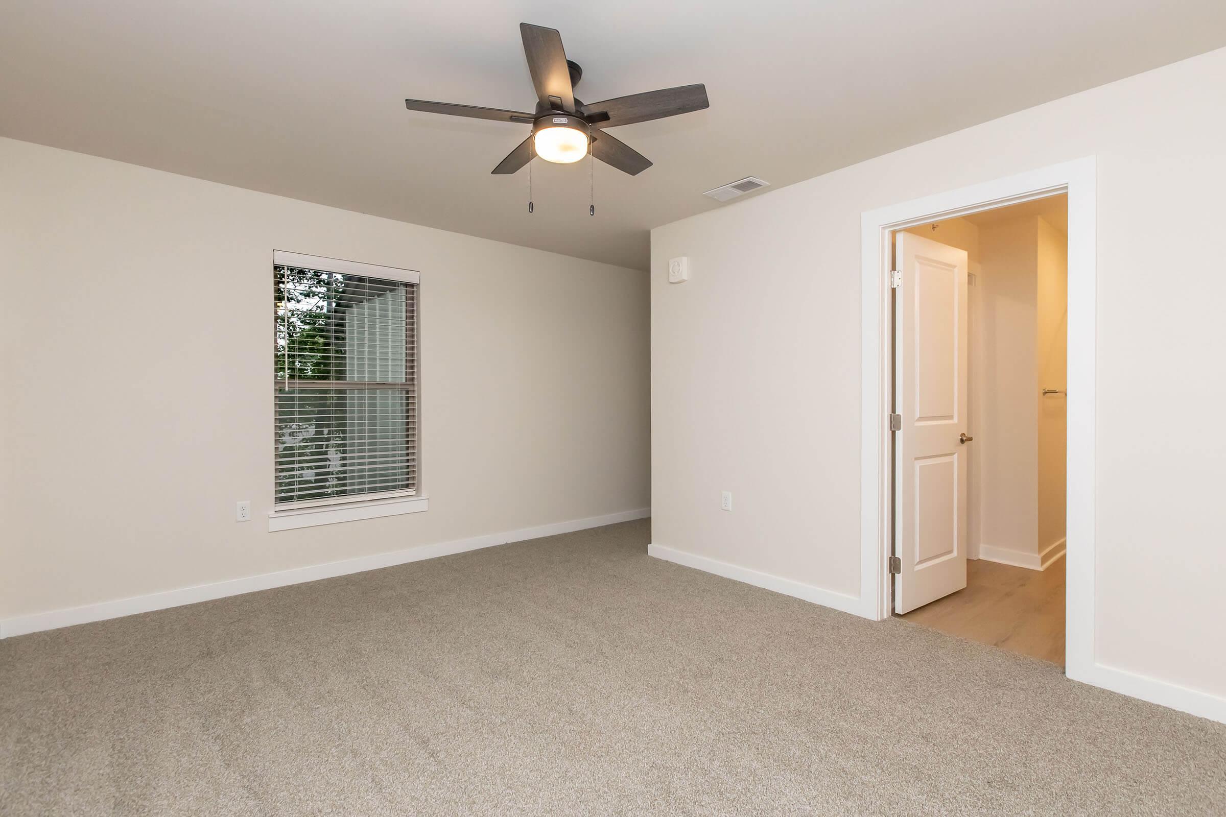 A spacious, empty room featuring a light ceiling fan, beige walls, and a large window with blinds. The floor is covered in light gray carpet, and there is a door leading to an adjacent room. Natural light fills the space, creating an inviting atmosphere.