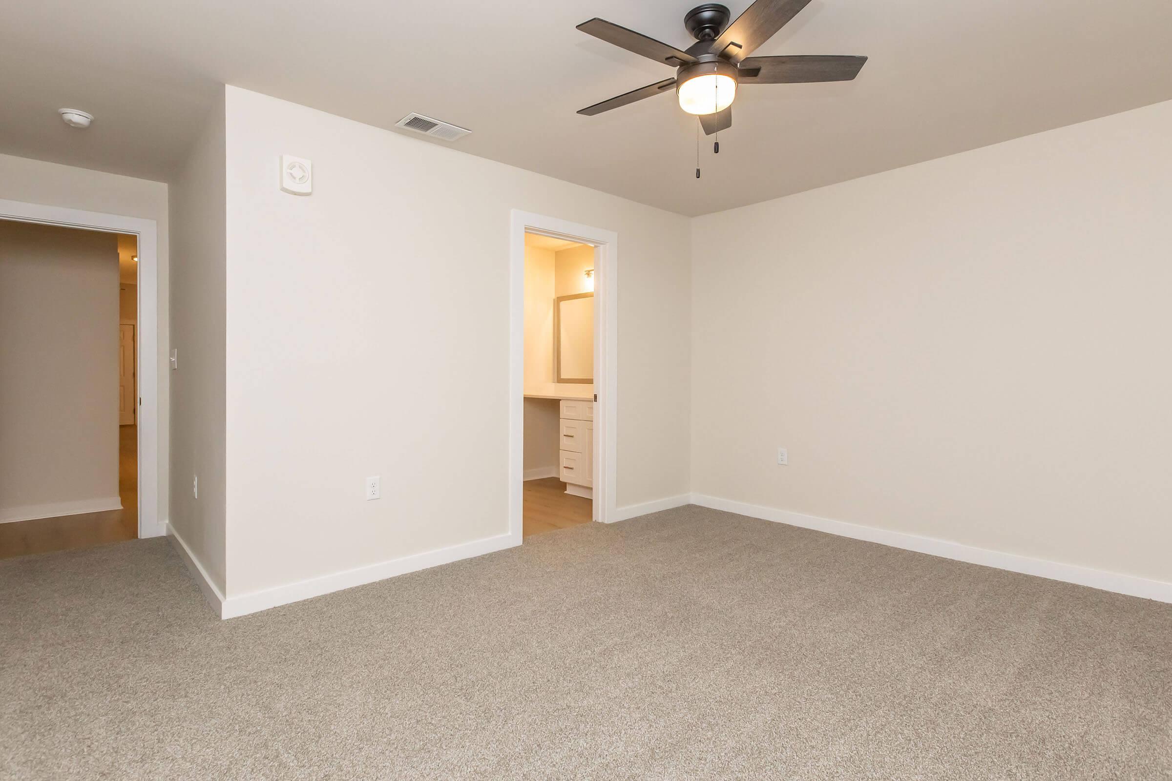 A spacious room with light beige walls and plush carpet flooring. It features a ceiling fan with wooden blades, a door leading to a bathroom area, and another room entrance visible in the background. The overall lighting is bright and inviting, creating a clean and minimalist aesthetic.