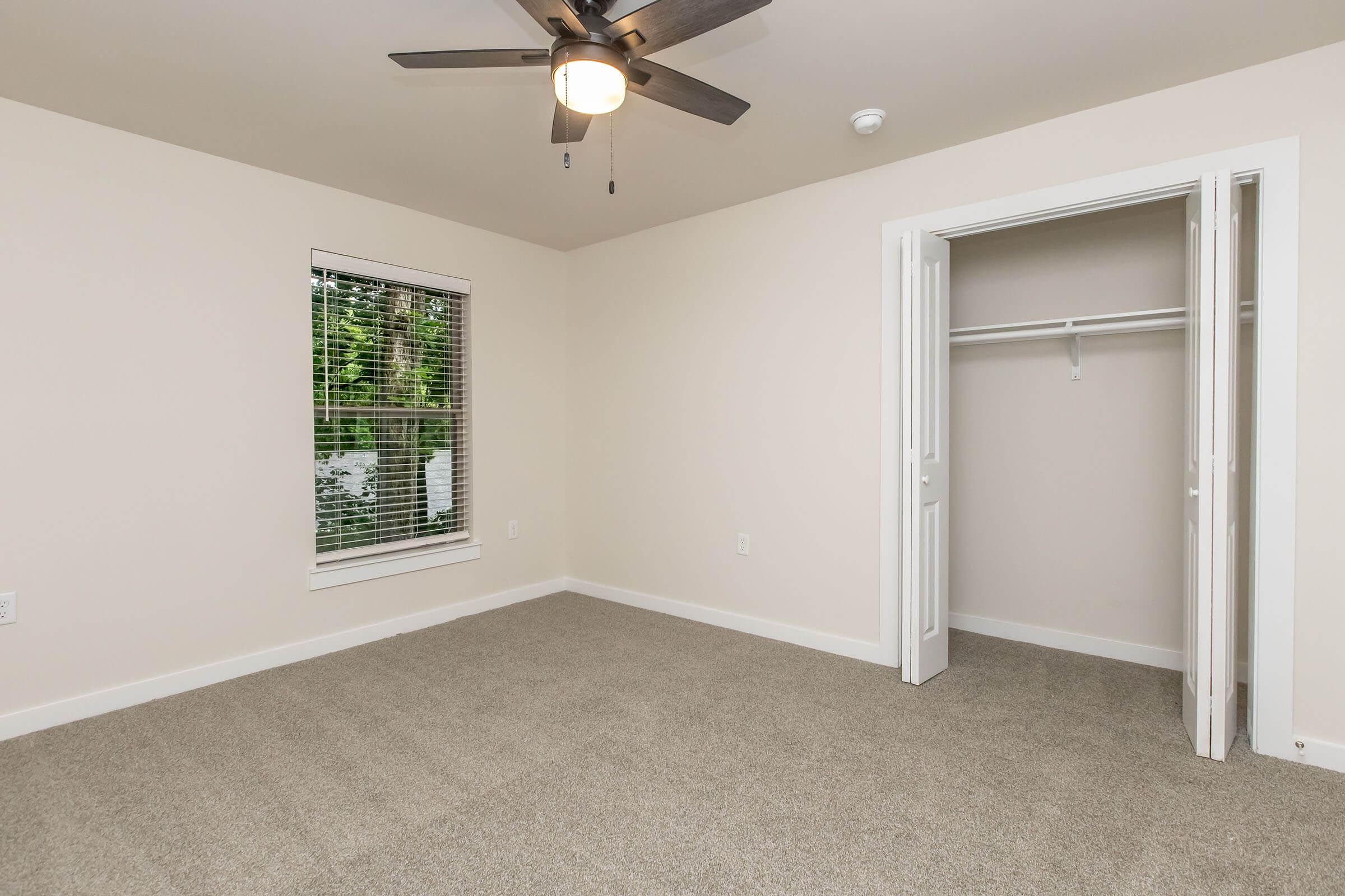 A spacious, empty room featuring a ceiling fan, light-colored walls, a window with blinds, and a built-in closet with doors slightly open. The flooring is carpeted in a neutral color, creating a clean and inviting atmosphere.