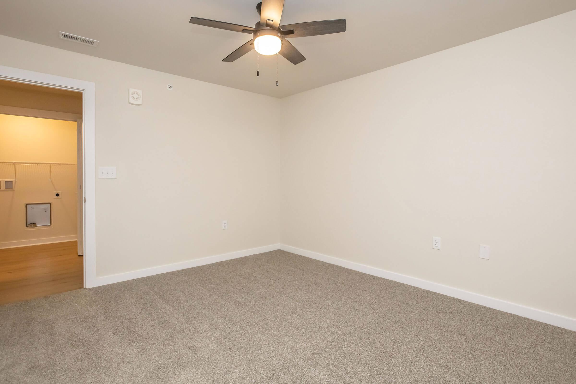 A minimalist room featuring light beige walls and plush gray carpeting. The ceiling has a ceiling fan with wooden blades. A doorway on the left leads to another area, and there is a wall socket visible. The space is empty, creating a clean and open atmosphere.