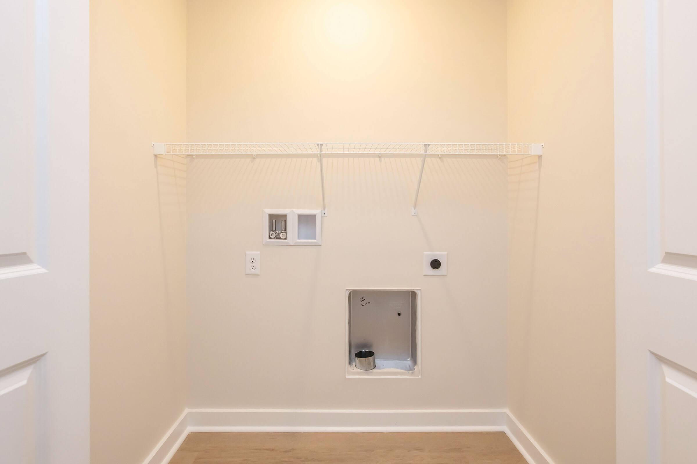 Empty laundry room with beige walls and a light-colored floor. A wire shelf is mounted on the wall, and there are electrical and plumbing connections visible. No appliances or items are present in the space, which is well-lit and organized.
