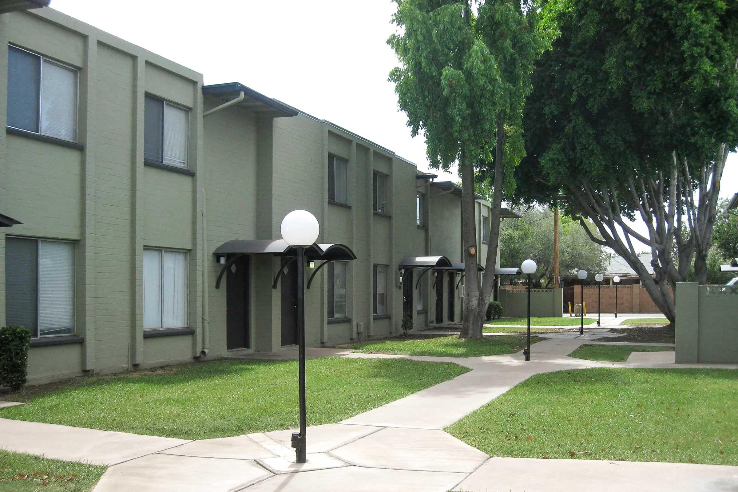 A view of a residential apartment community featuring several green-painted buildings. The pathway is lined with neatly trimmed grass and lampposts, providing a well-maintained outdoor space. Lush trees can be seen in the background, contributing to a serene atmosphere.