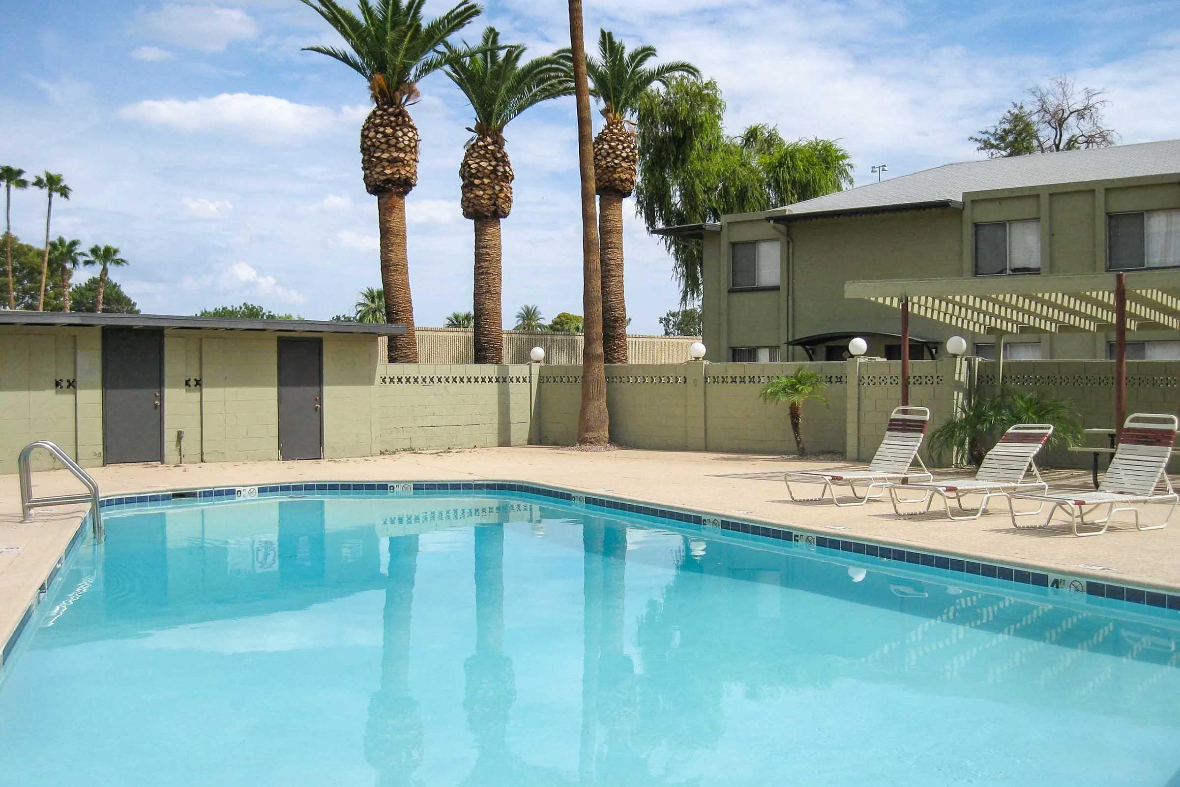 A serene outdoor swimming pool surrounded by palm trees and lounge chairs. The water is clear and inviting, with a light blue tile border. A few buildings can be seen in the background, under a partly cloudy sky, creating a relaxing atmosphere in a sunny setting.