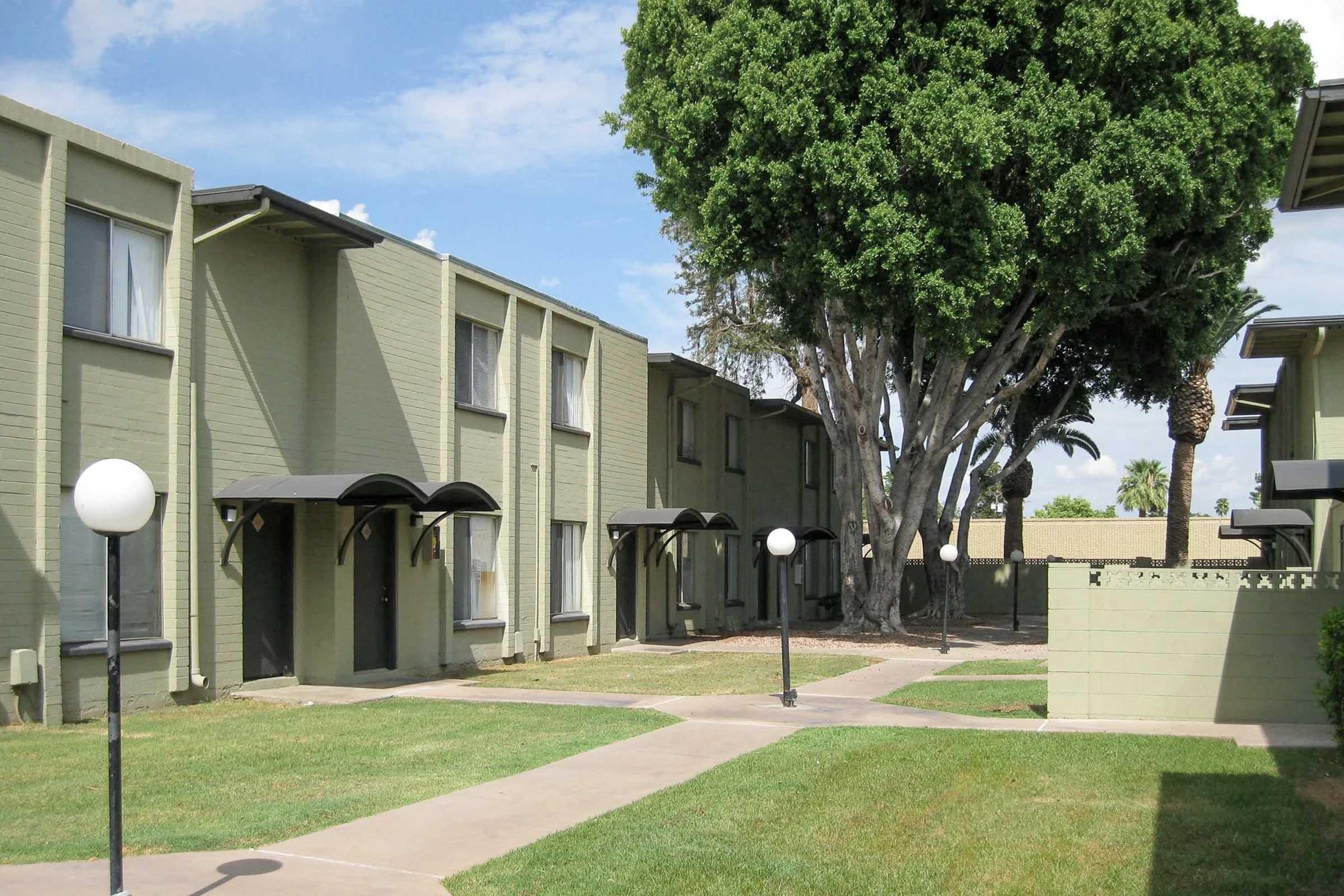 A row of green apartment buildings with arched doorways is visible. In the foreground, a well-maintained lawn features round white light fixtures along a pathway leading through the complex. A large tree provides shade, with blue skies and scattered clouds visible above.