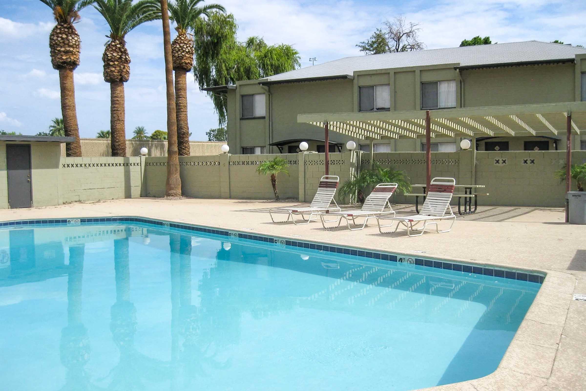 Swimming pool with clear blue water surrounded by a concrete deck. There are several lounge chairs placed around the pool, palm trees in the background, and a green building with a covered patio area. The scene is bright and sunny, conveying a relaxed outdoor atmosphere.