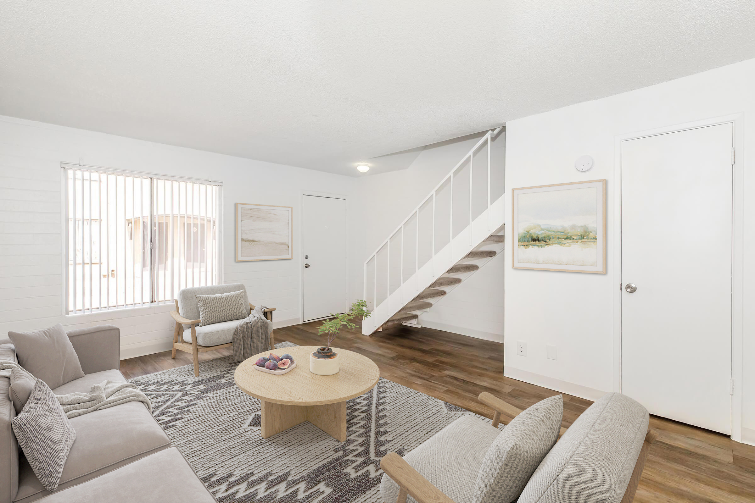 A bright, modern living room featuring a cozy seating area with light-colored sofas, a round wooden coffee table, and a decorative rug. The walls are white, with a staircase visible leading to an upper level. Natural light streams in through a window with blinds. A framed artwork is displayed on the wall.