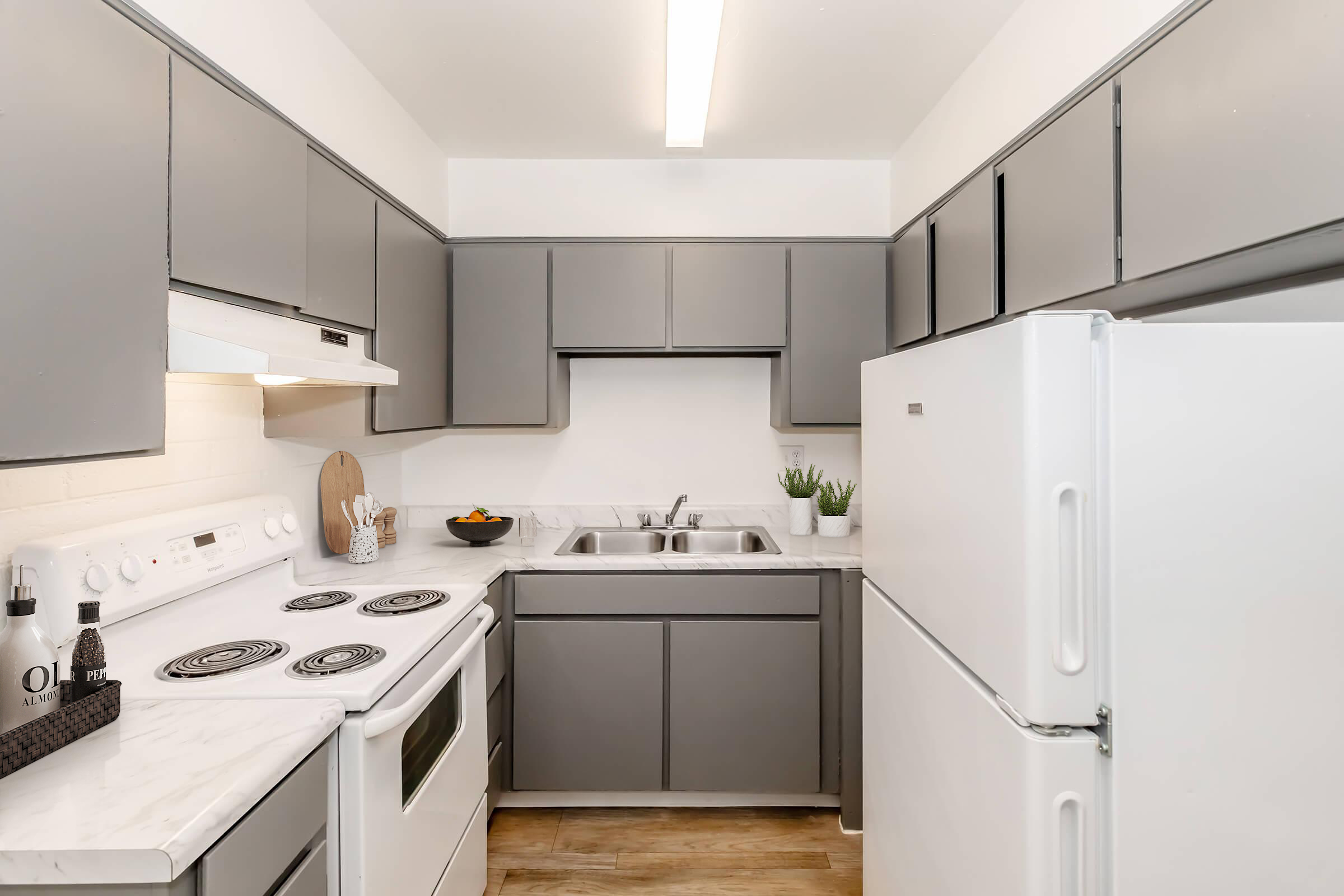 A modern kitchen featuring gray cabinets, a white refrigerator, and a marble countertop. The space includes a stove and oven with four burners, a double sink, and a small bowl of fruit on the counter. There are also decorative plants and kitchen accessories for a cozy atmosphere.